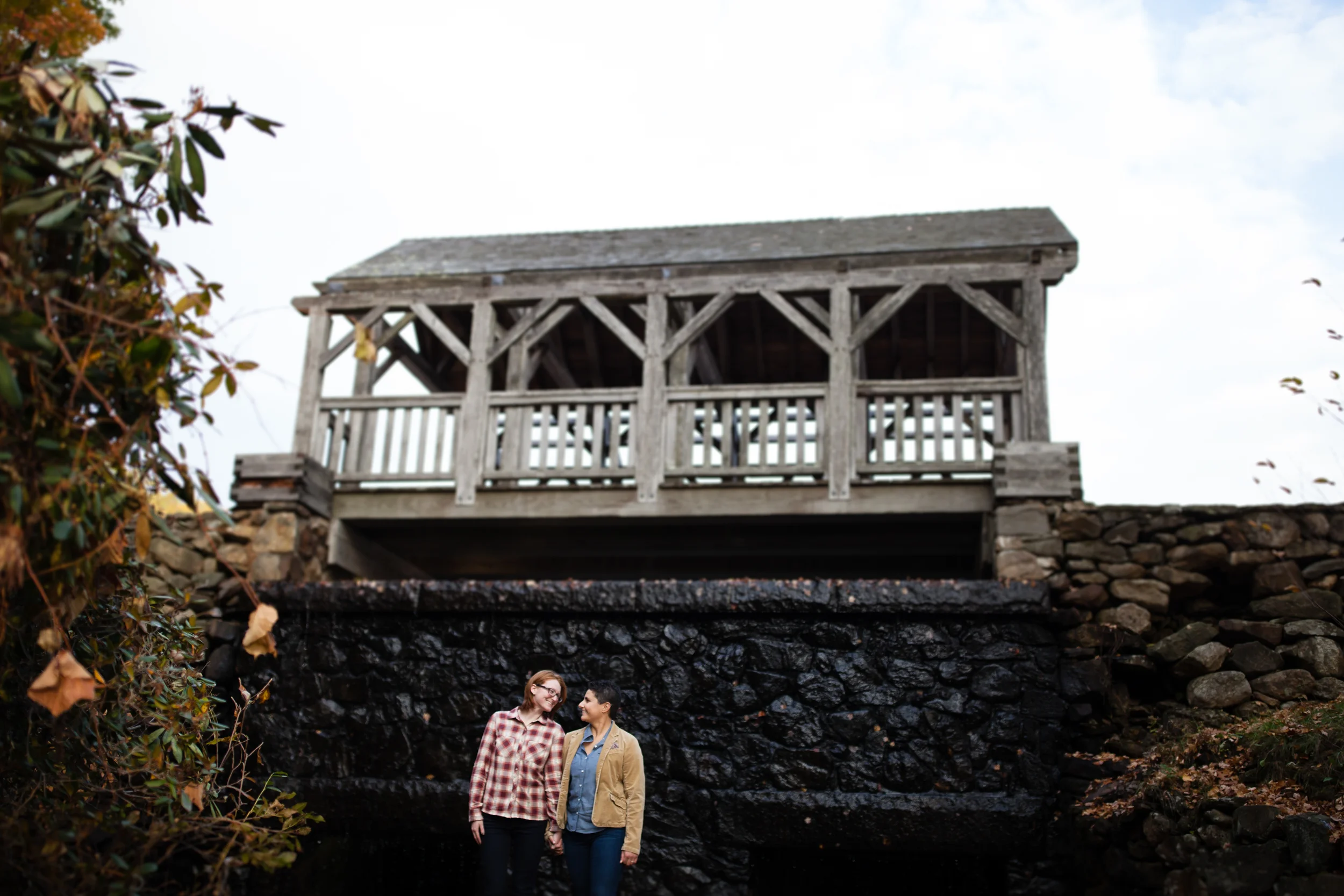 Two women standing close together and smiling, near a small black waterfall with a wooden structure above them.