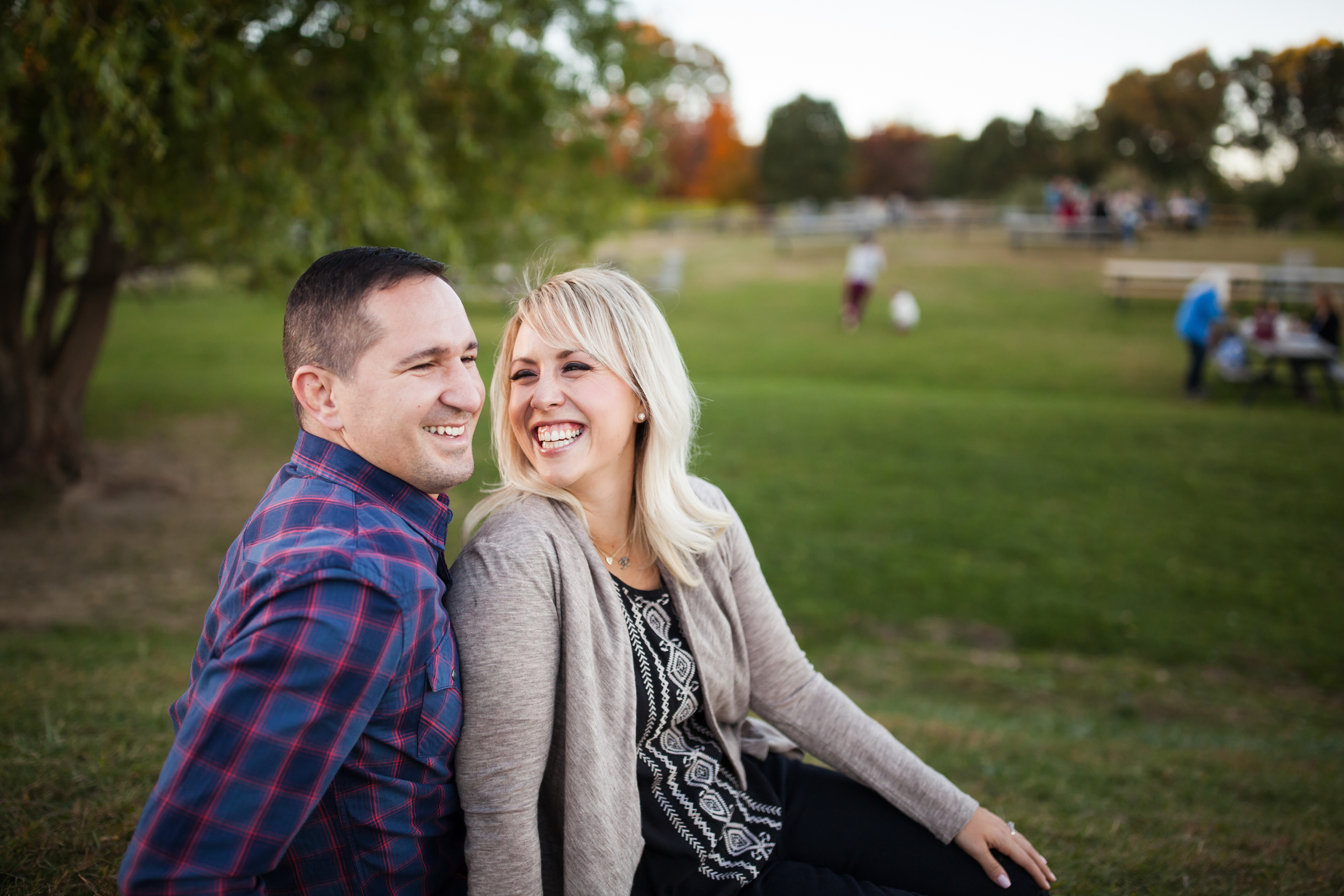 A couple sitting on the grass in a park, smiling and laughing together, with trees and people walking or sitting at picnic tables in the background.