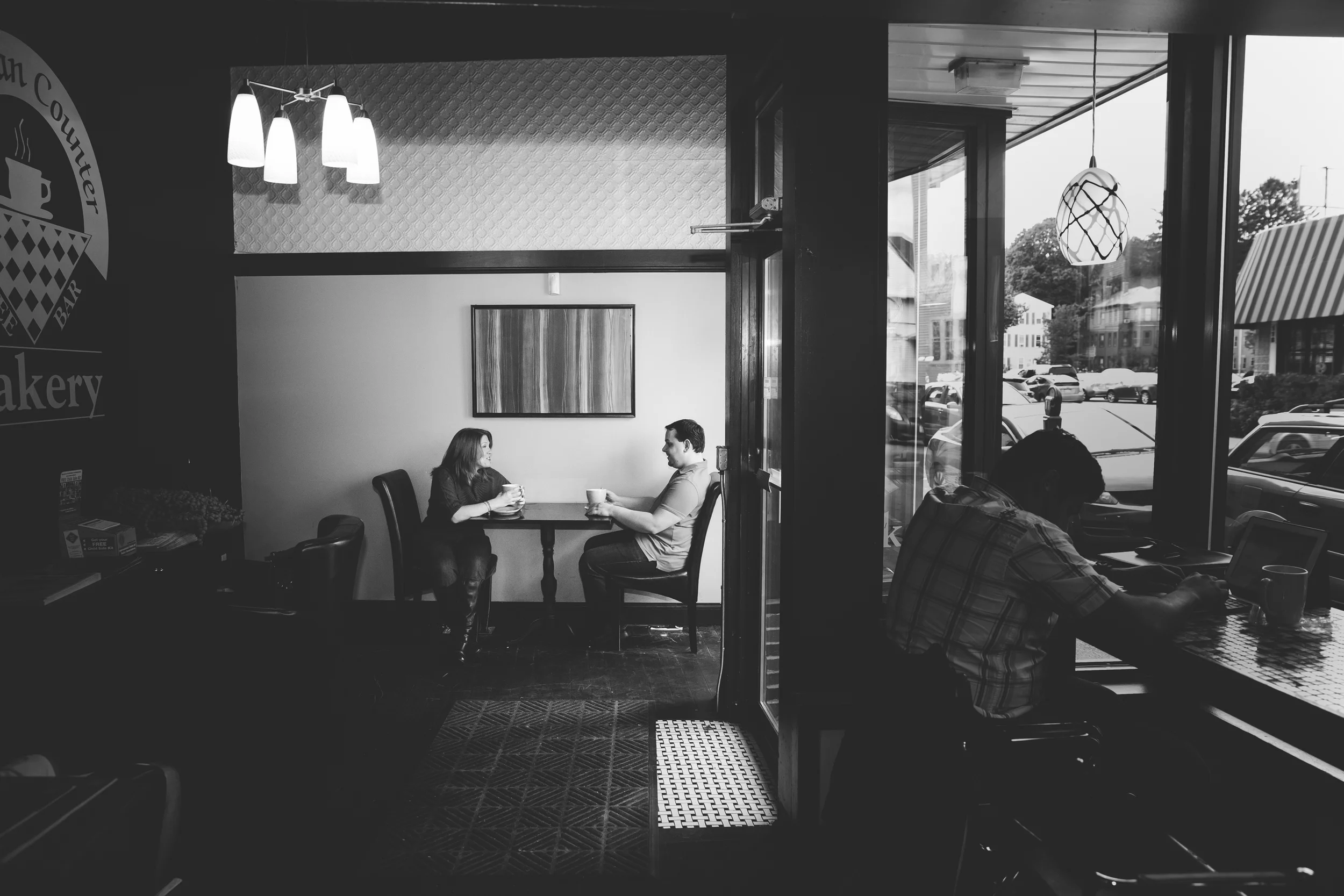 Inside a coffee shop with two people sitting at a table by the window, a woman and a man, drinking coffee and talking. Another person is working on a laptop near the window. Outside the window, parked cars and buildings are visible.