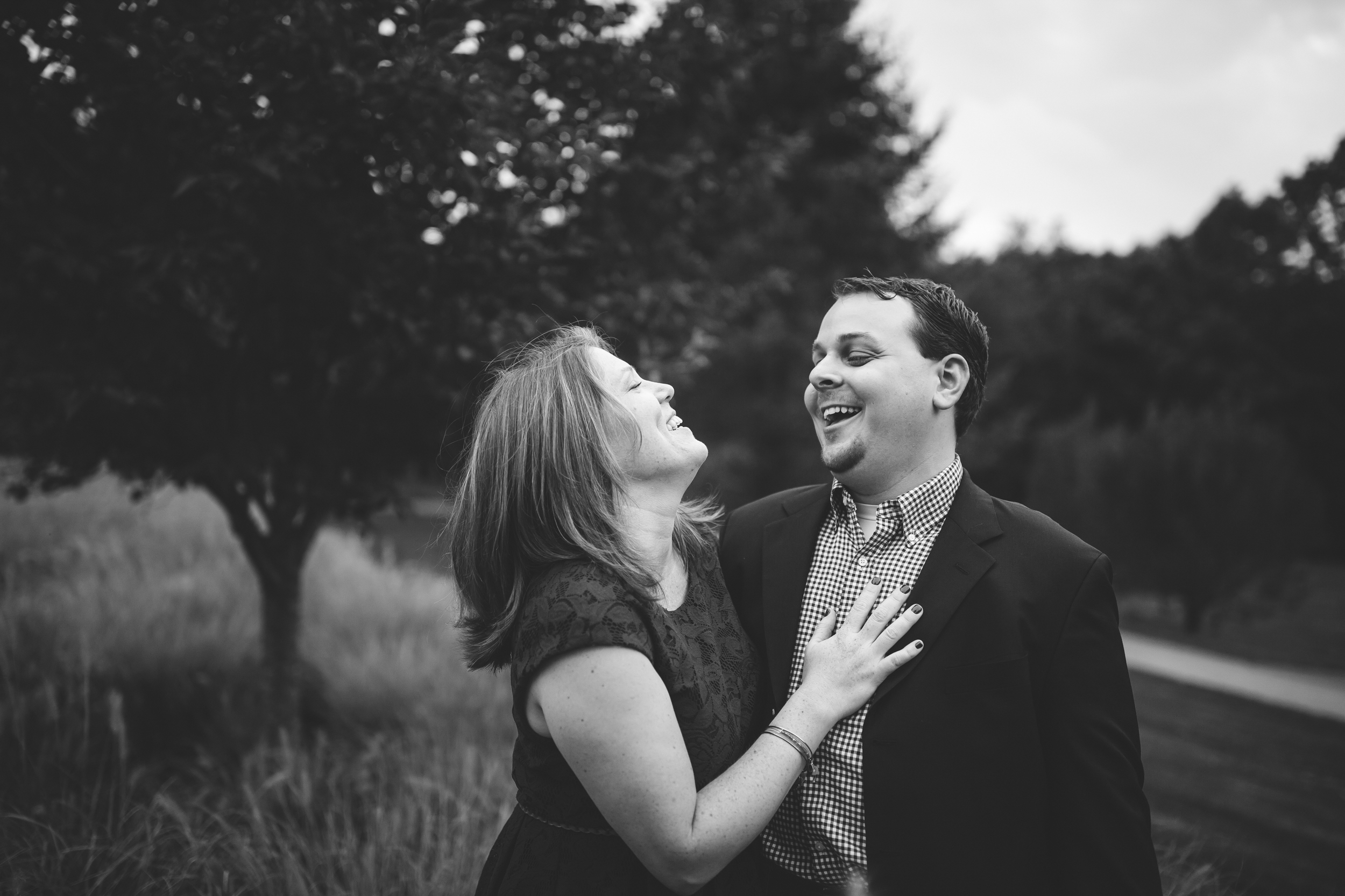 A black and white photo of a happy couple outdoors, smiling and laughing as they look at each other. The woman has shoulder-length hair and is wearing a dress, while the man is dressed in a suit with a checkered shirt. They are standing in a grassy a