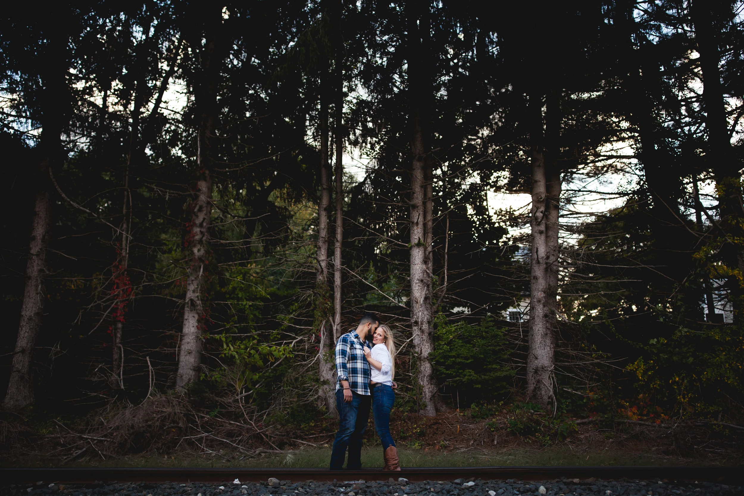 A couple standing close together on a railroad track in front of a dense forest at dusk.
