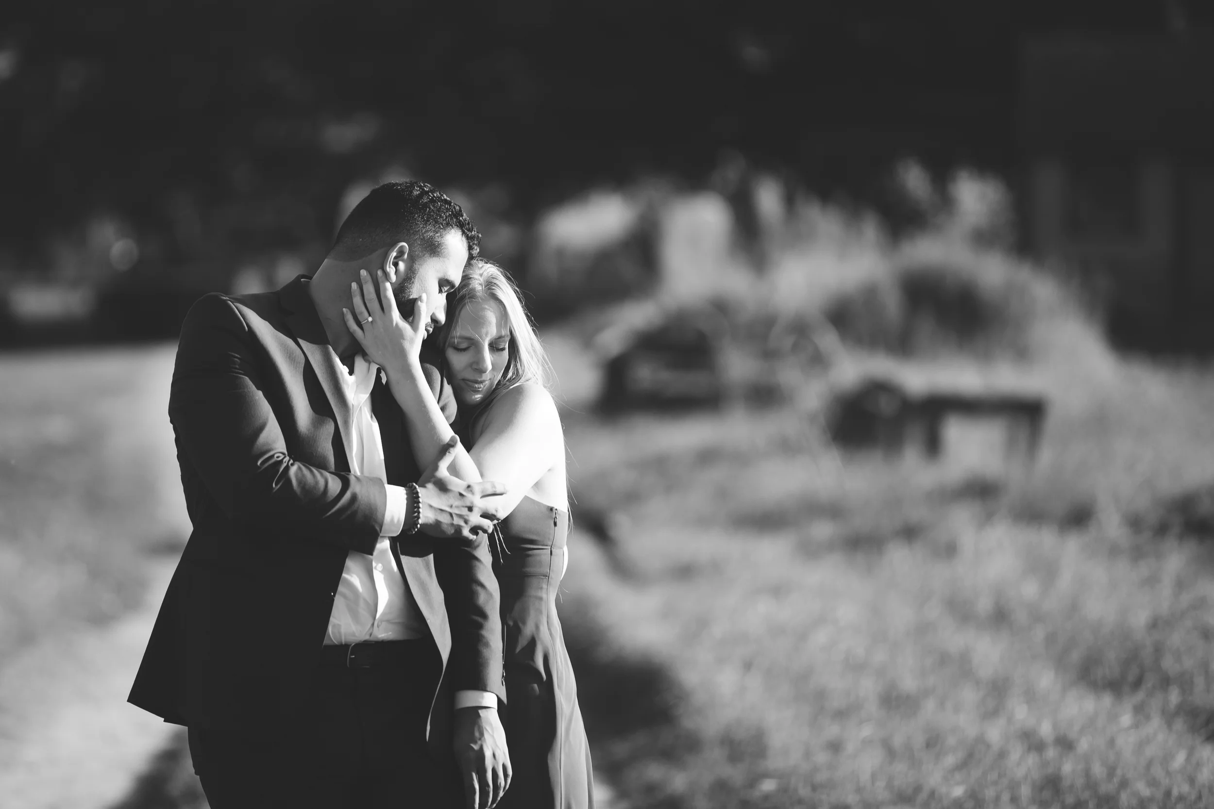 A black and white photo of a couple standing outdoors, closely embracing and looking at a phone, with a blurred background of park benches and trees.