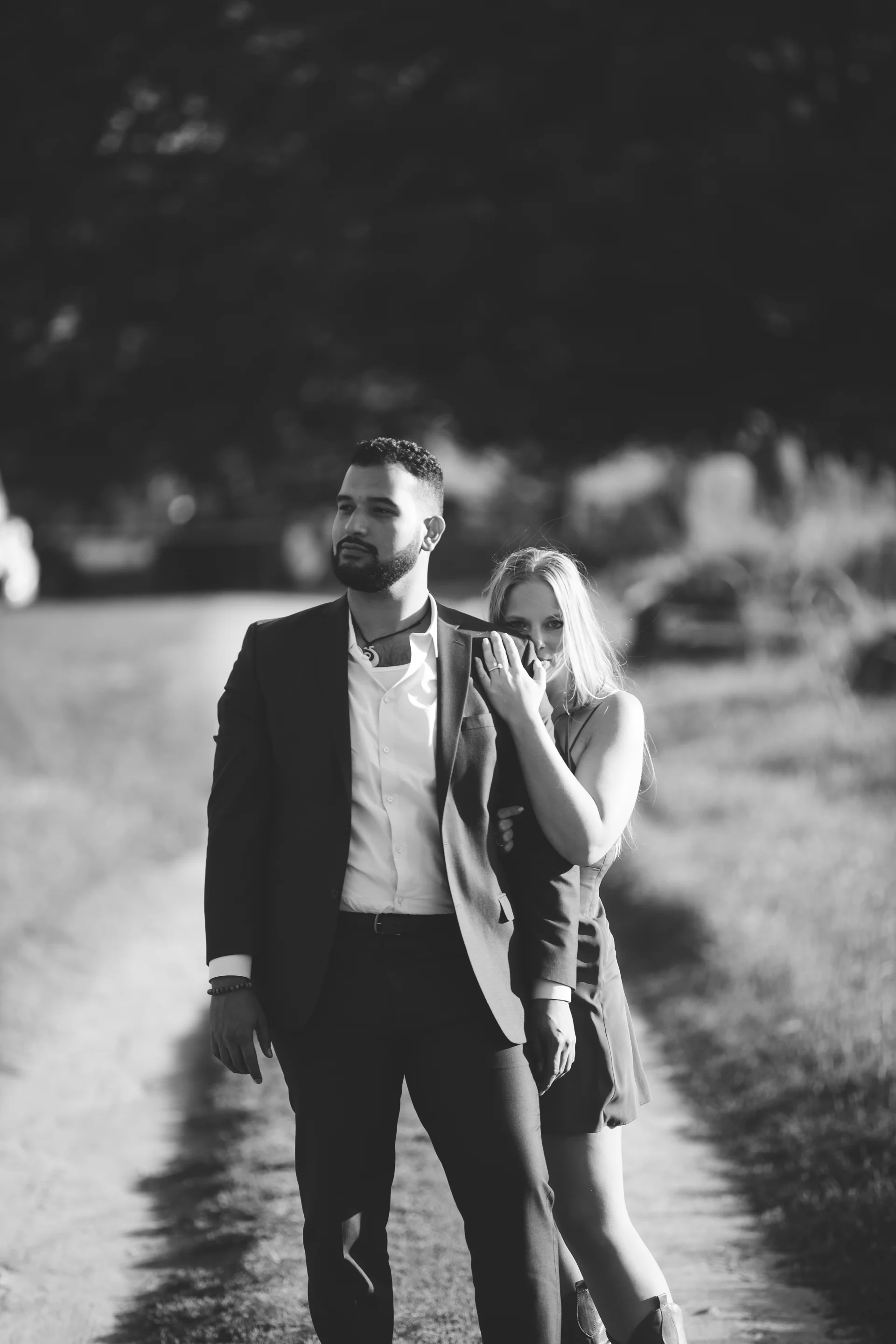 Black and white photo of a man in a suit and a woman in a dress standing outdoors on a dirt path with trees in the background.