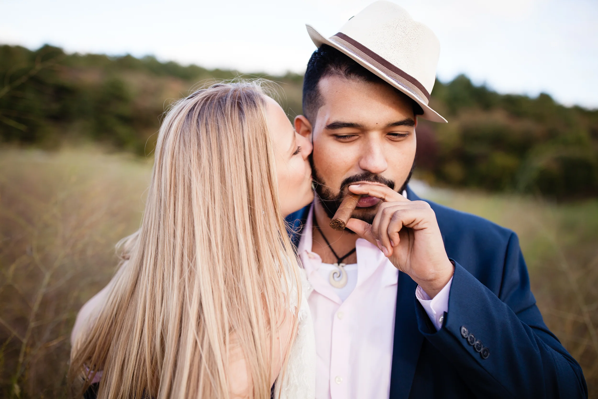 A woman is whispering to a man holding a cigar, who is wearing a suit and a fedora hat outdoors with a blurred natural background.