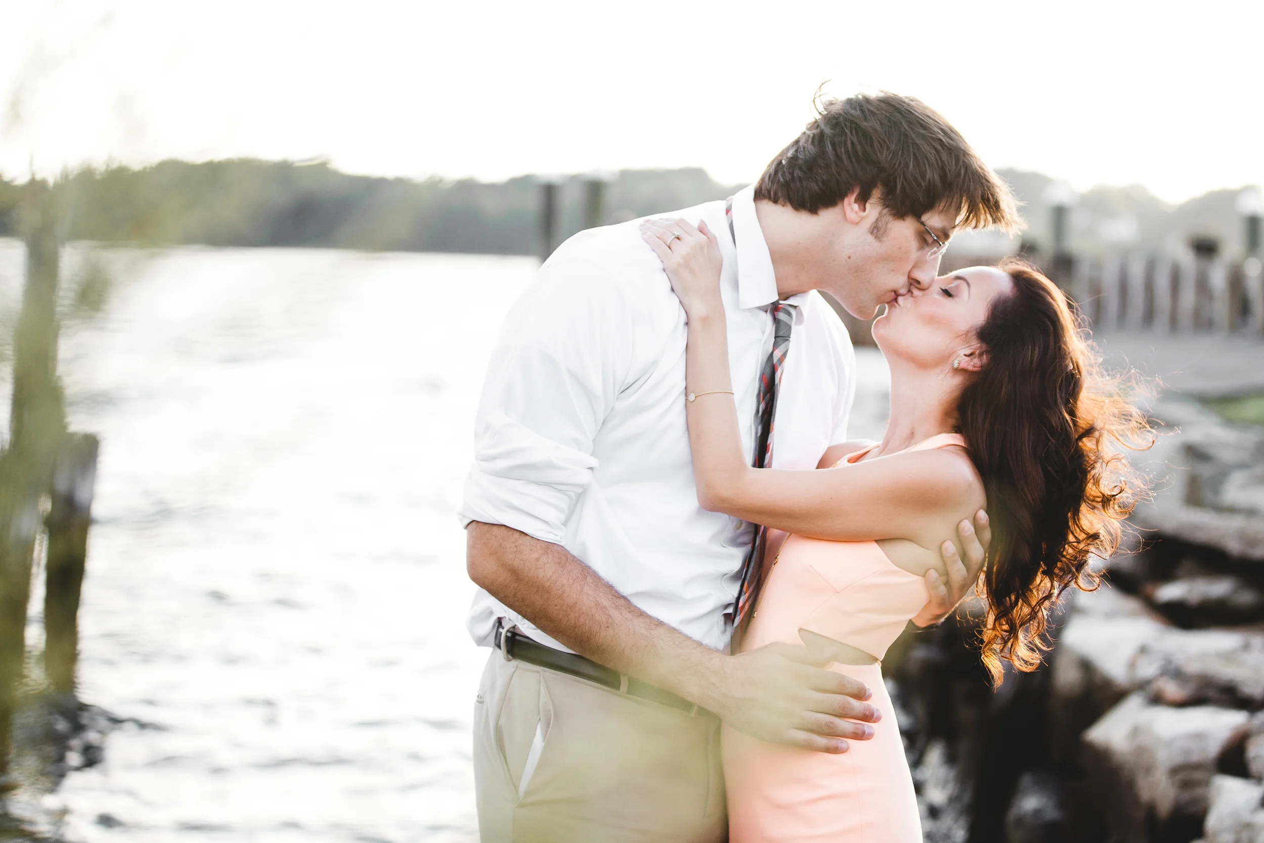 A couple kissing by the water at sunset, with the man wearing a white shirt and tie, and the woman in a peach dress, embracing each other lovingly.