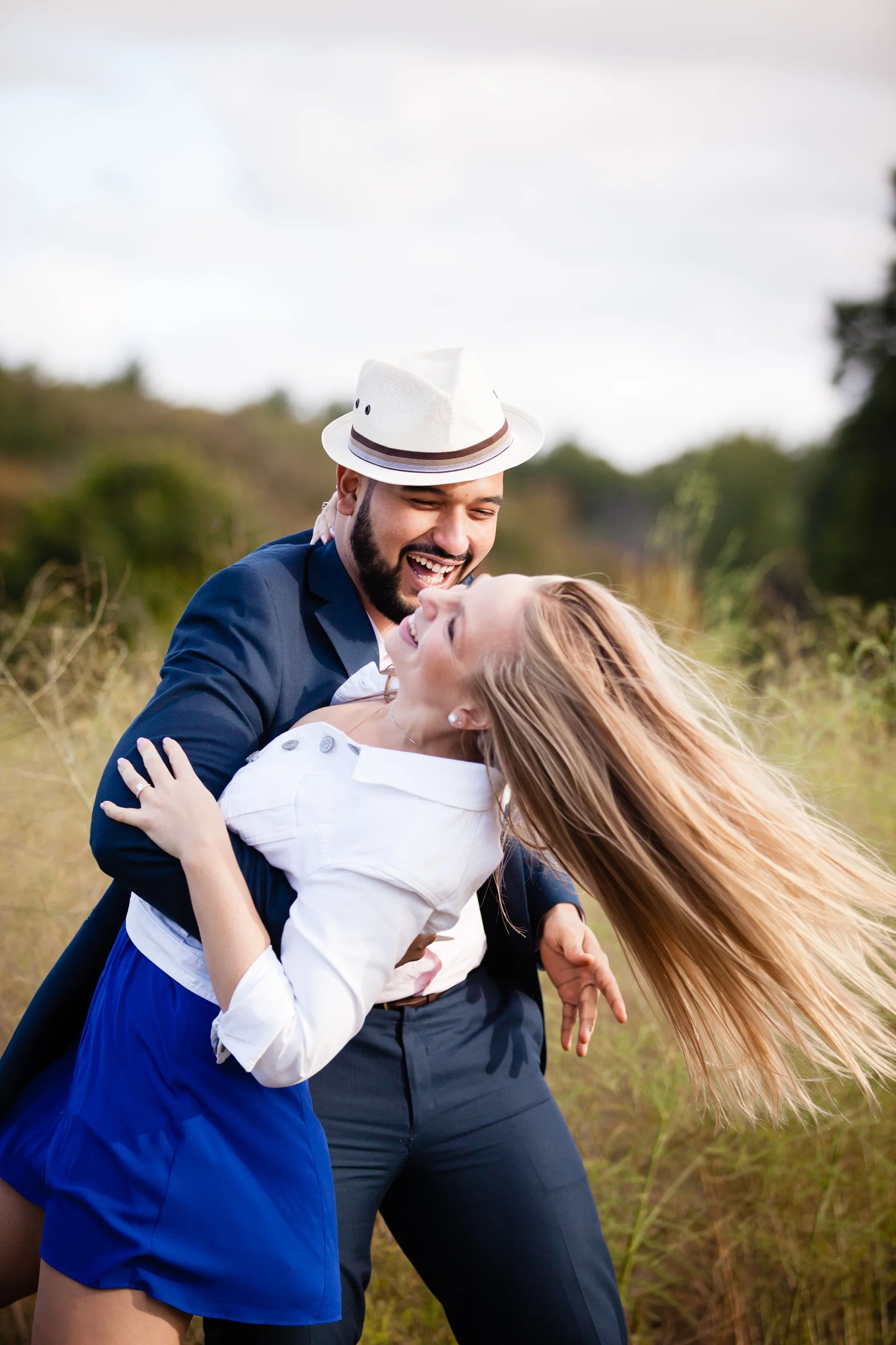 A man and woman are dancing and smiling outdoors in a grassy field with a cloudy sky in the background.
