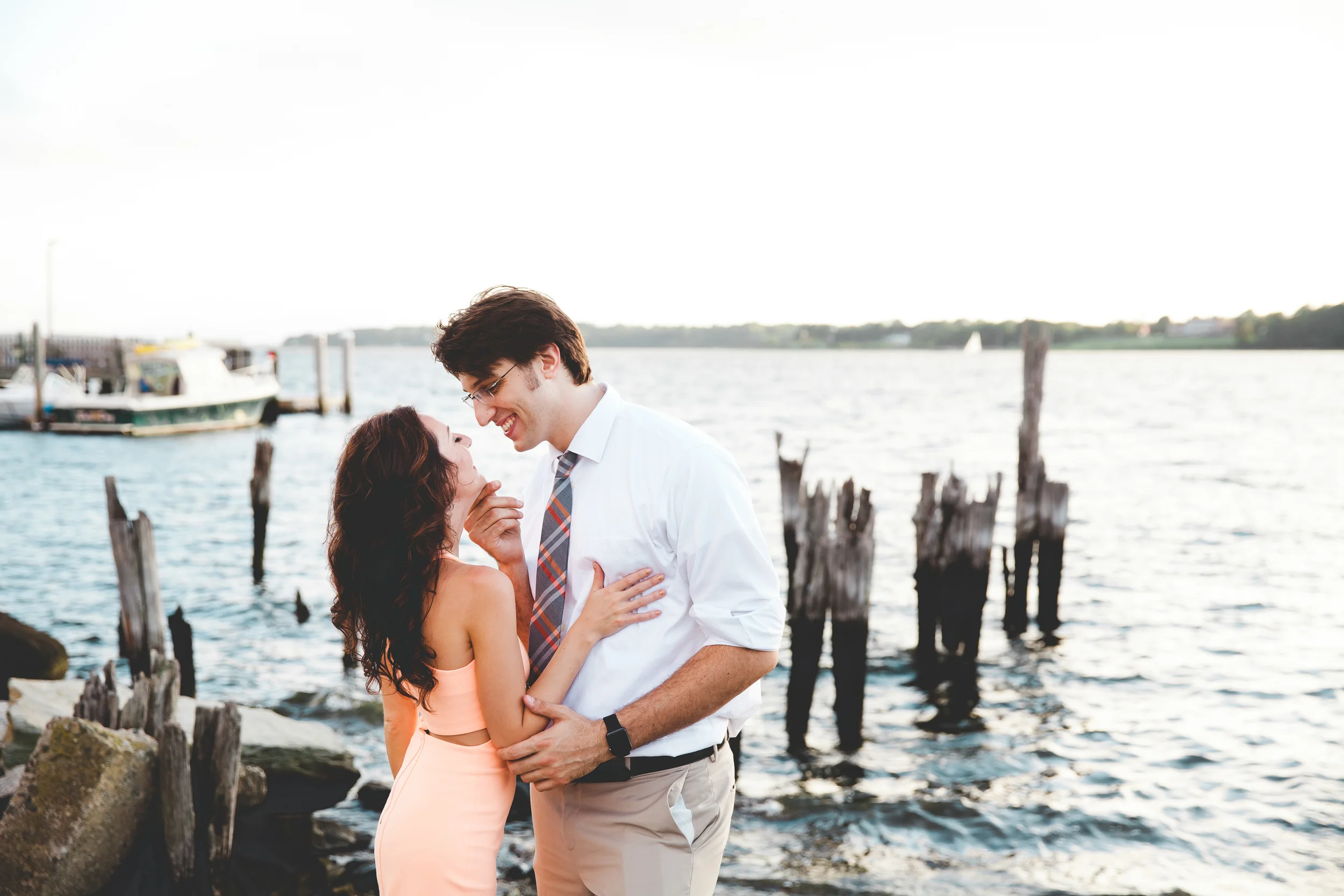 A young couple smiling and looking at each other by the water, with a dock and boats in the background during the evening.