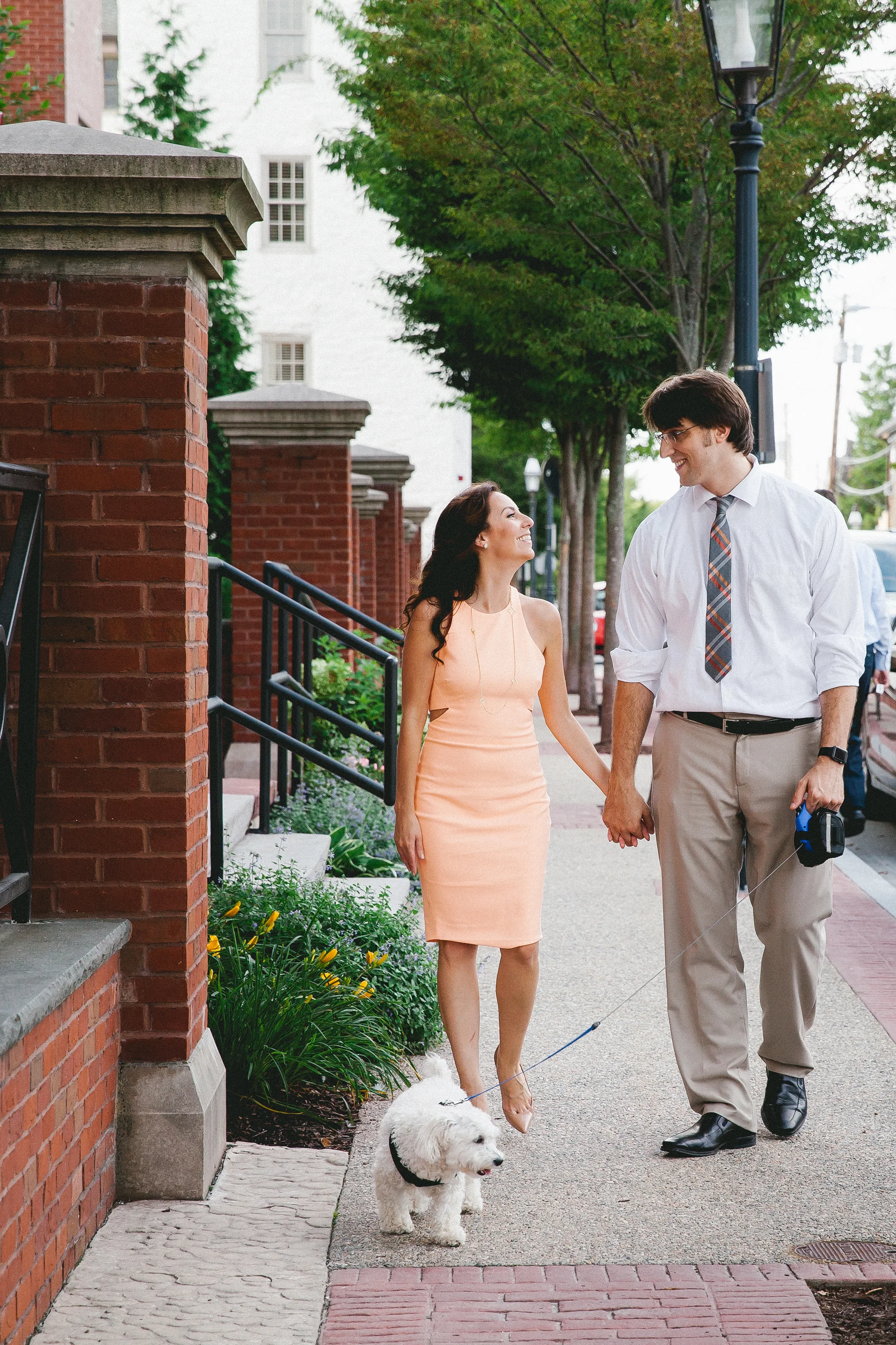 A woman and a man walking a small white dog on a city sidewalk, smiling at each other.