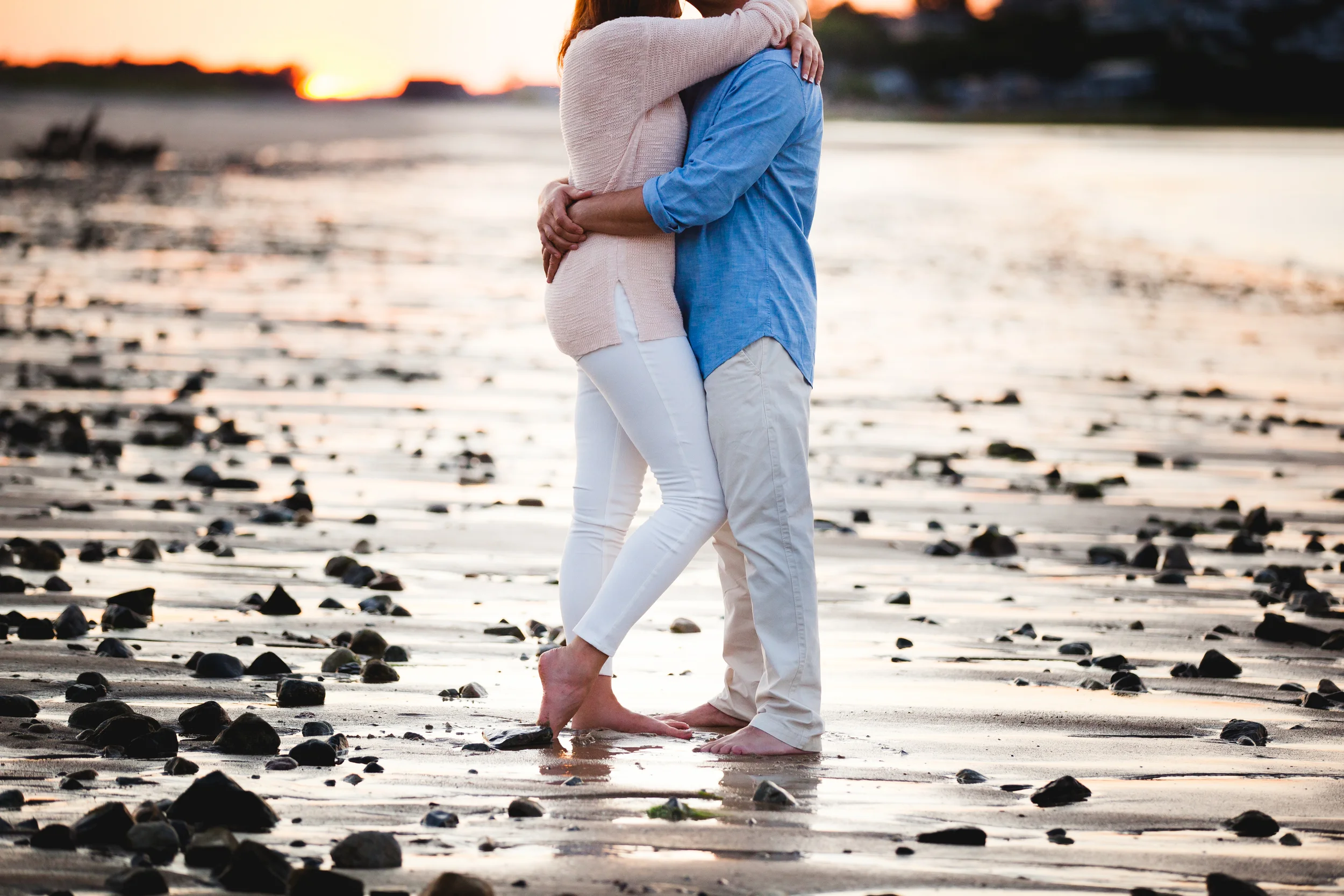 A couple embracing on a rocky beach during sunset, with their feet in the shallow water.