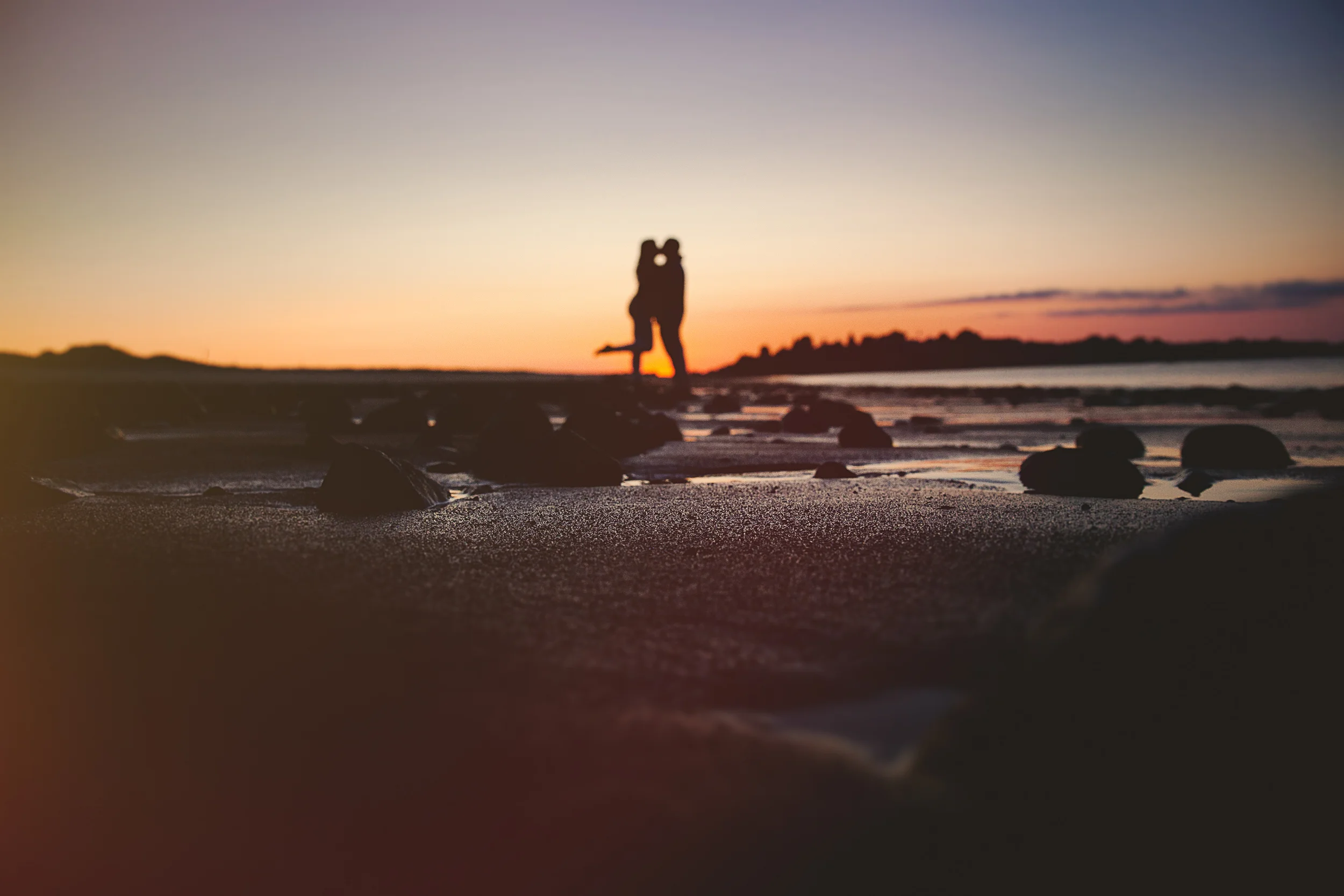 Silhouetted couple on a beach during sunset, with rocks on the sand and the ocean in the background.