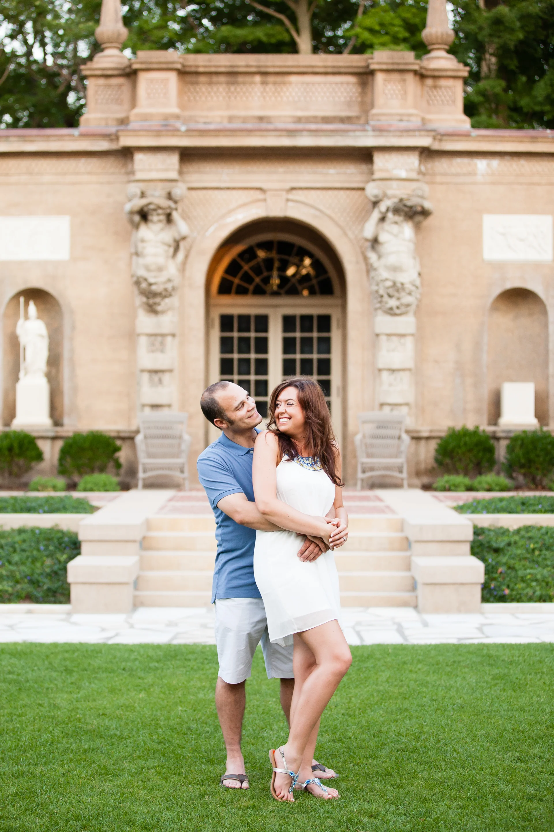 A couple embracing and smiling in front of a historic stone building with statues, steps, and greenery.