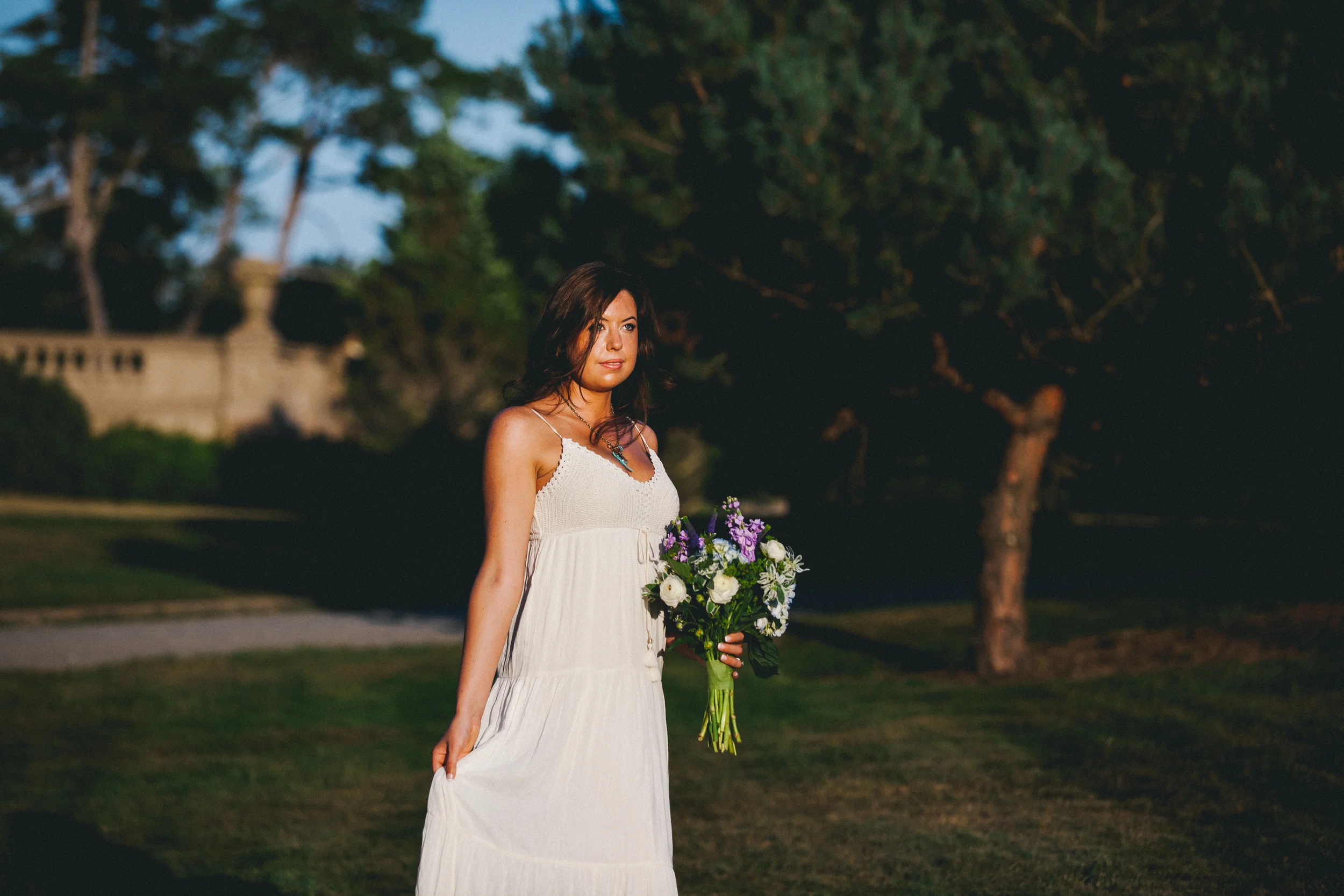 A woman in a white dress holding a bouquet of flowers outdoors during evening or sunset with trees and a stone fence in the background.