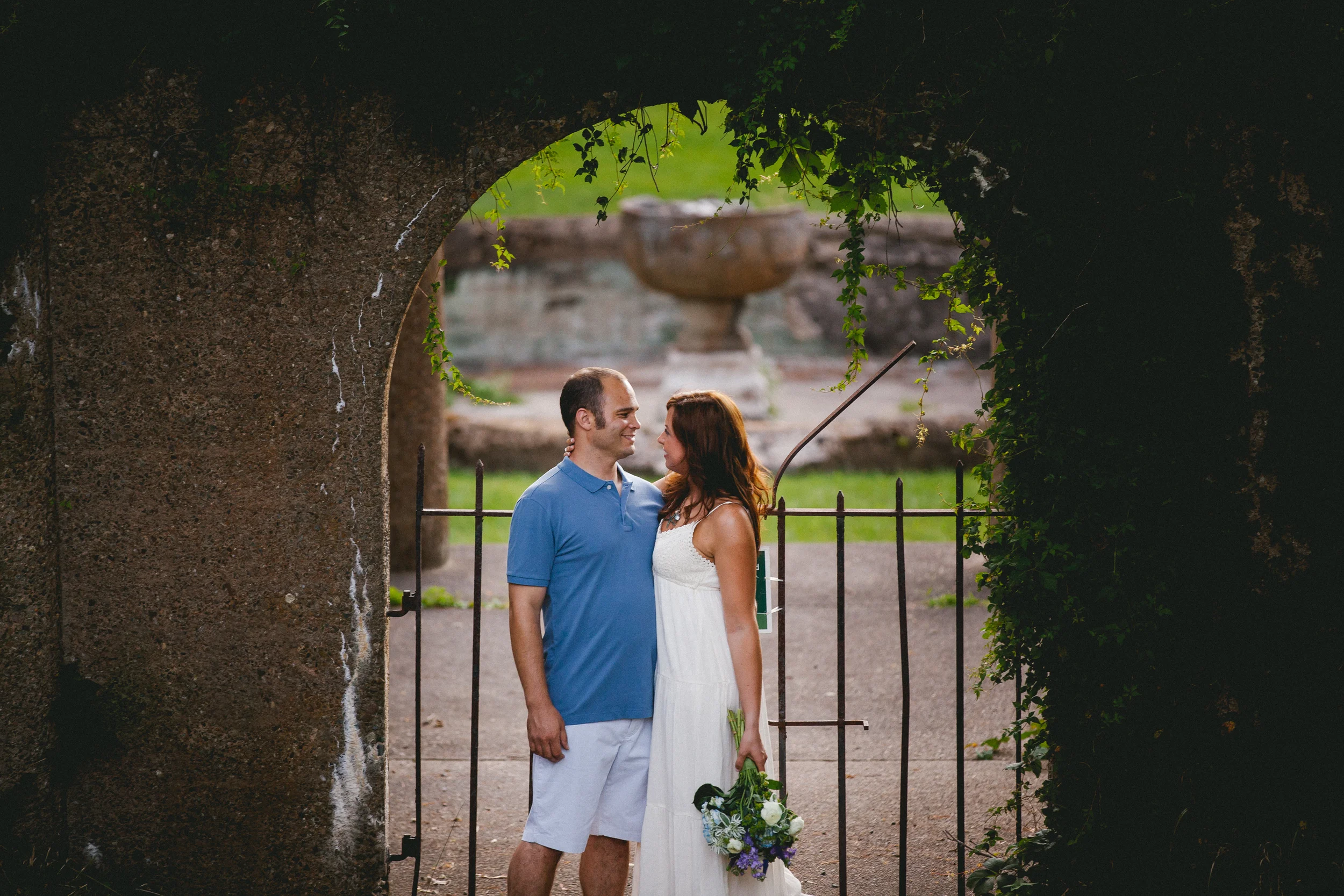 A couple stands close together, smiling, through an archway with green foliage, with a fountain in the background. The woman is holding a flower bouquet.