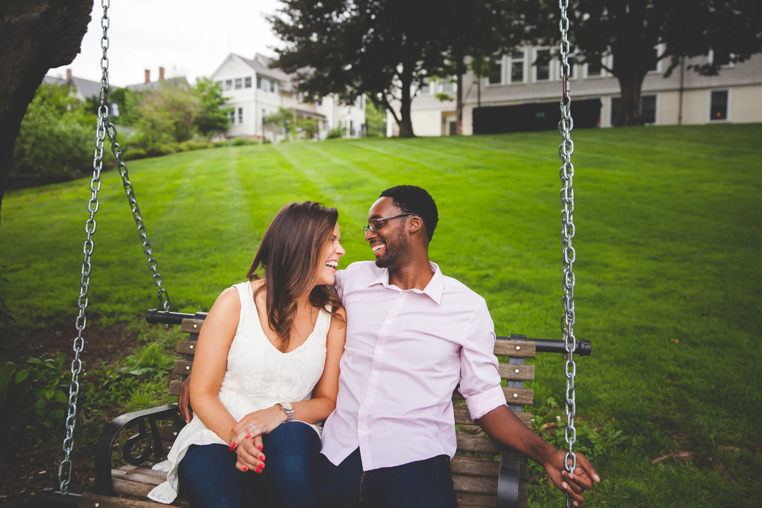A happy couple sitting together on a swing in a park, smiling and looking at each other, with green grass and houses in the background.