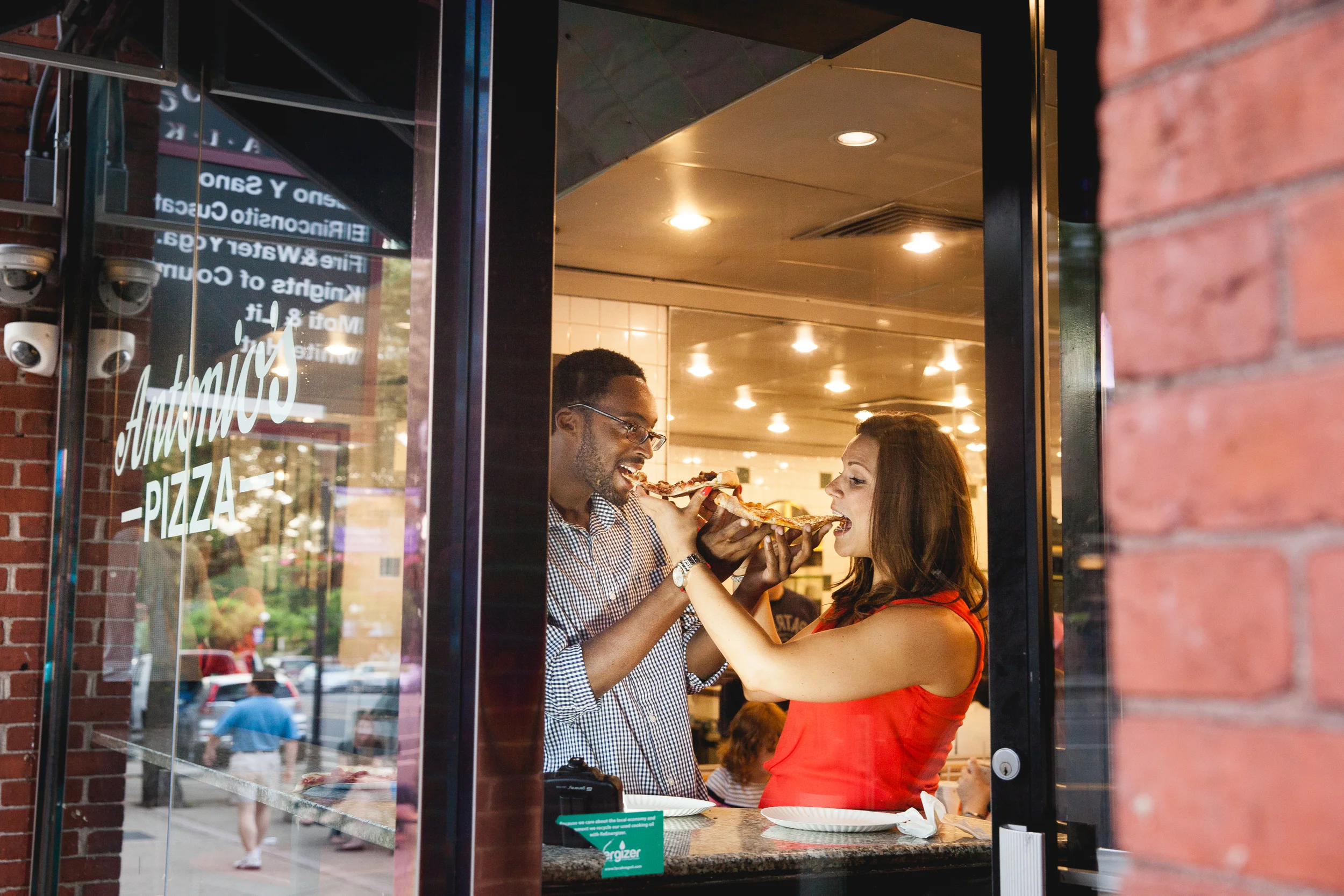 A man and woman sharing a slice of pizza inside a pizzeria viewed through a glass window with the pizzeria's name and description on the glass.