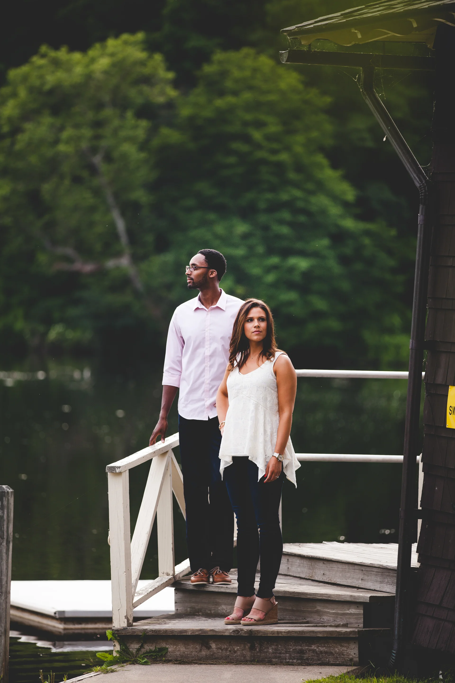 A man and woman stand on a small wooden dock near a body of water surrounded by green trees. The man, wearing glasses, a white shirt, and dark pants, is looking to the left, while the woman, in a white sleeveless top, dark jeans, and beige heels, is 
