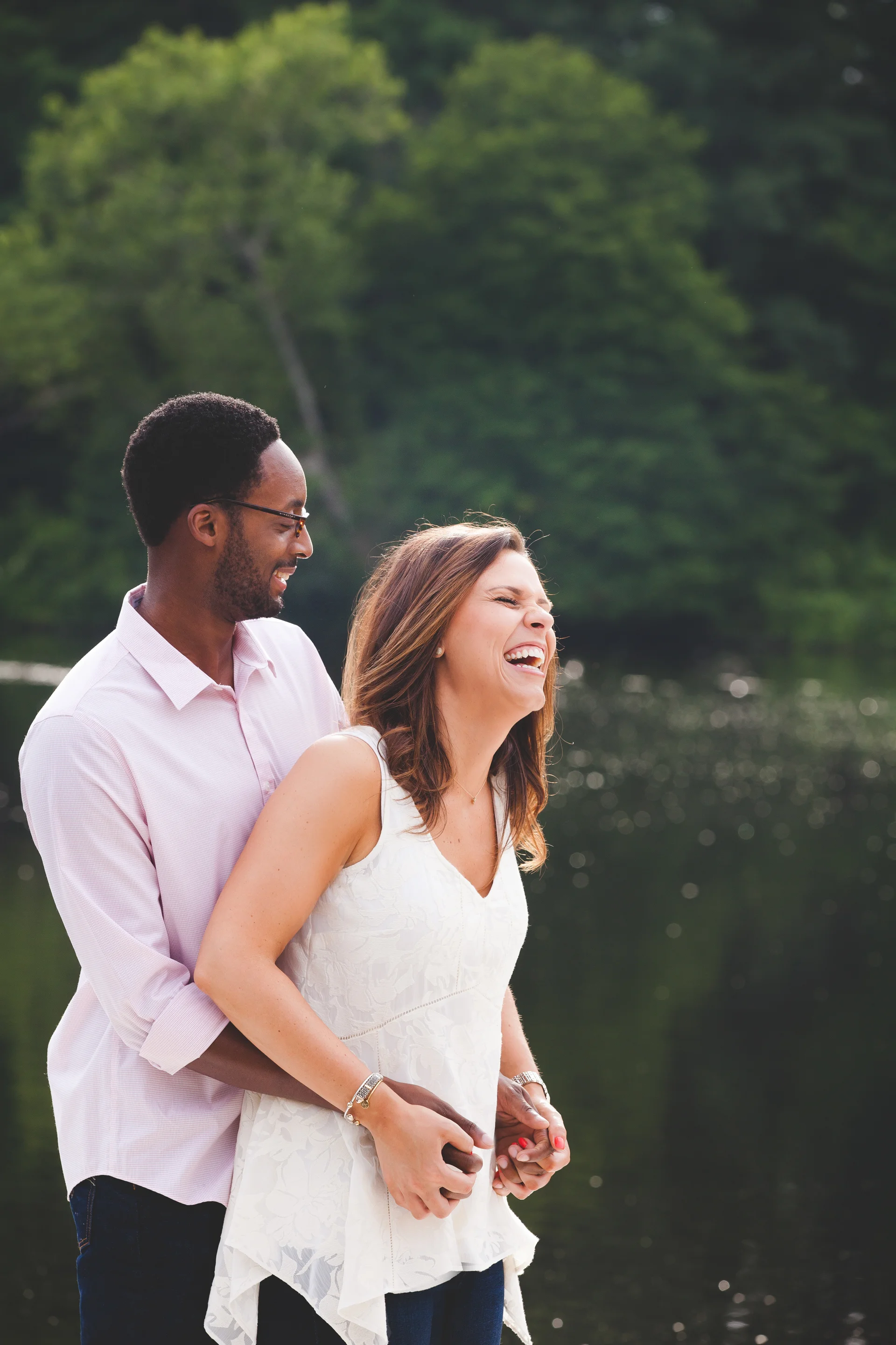 A couple laughing and embracing outdoors near a body of water with a green forest background.