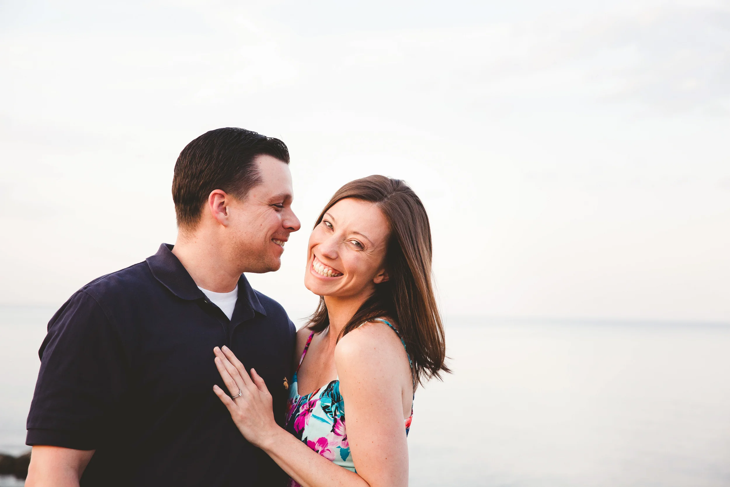 A happy couple standing close together on a beach, smiling, with the ocean and sky in the background.