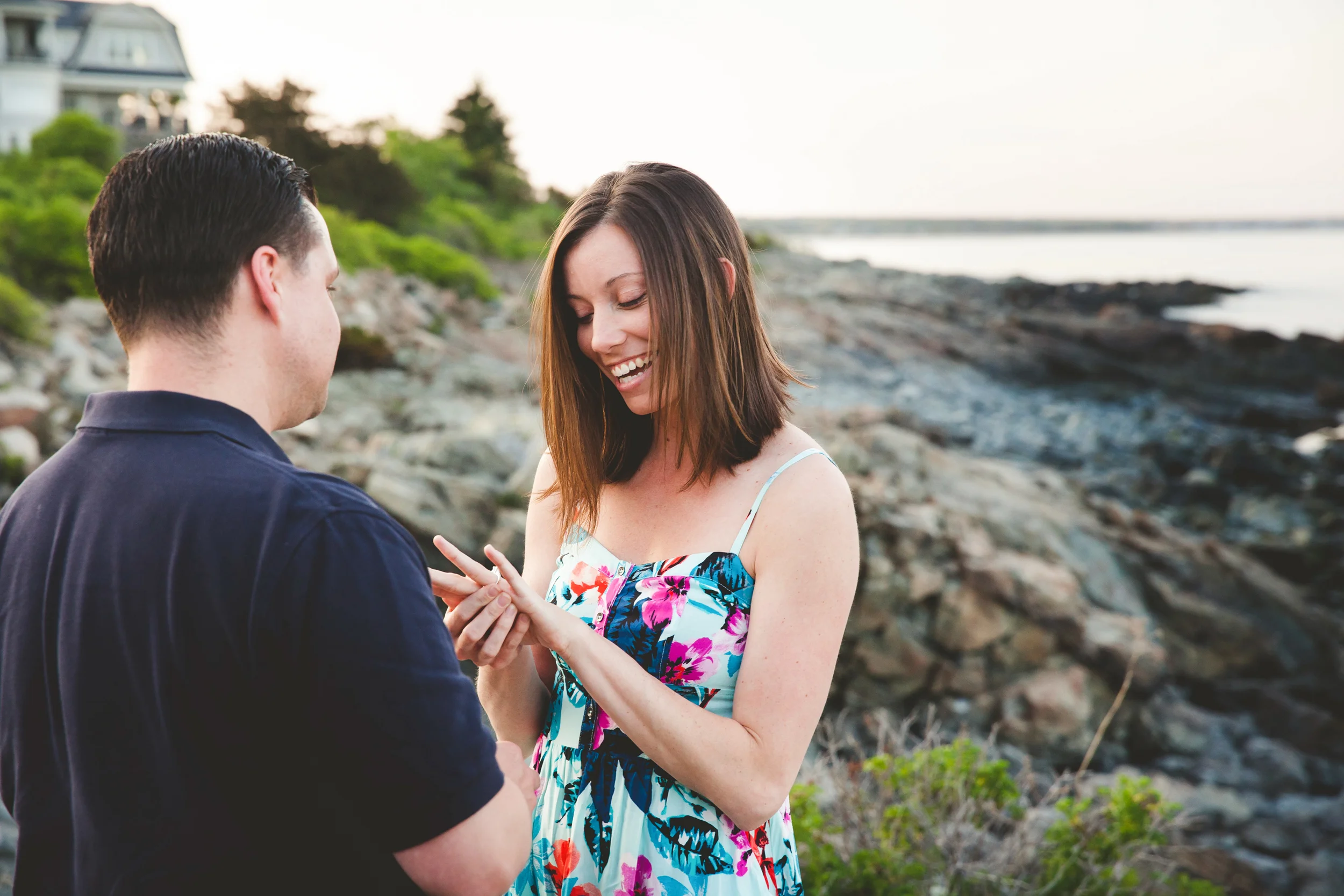 A woman is smiling as a man puts a ring on her finger outdoors near the water with rocks and greenery, during sunset.