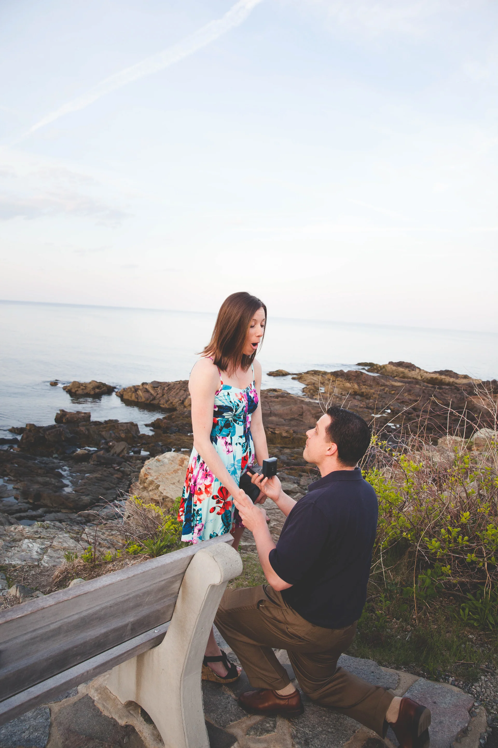A man proposing marriage to a woman on a rocky beach by the water during sunset.