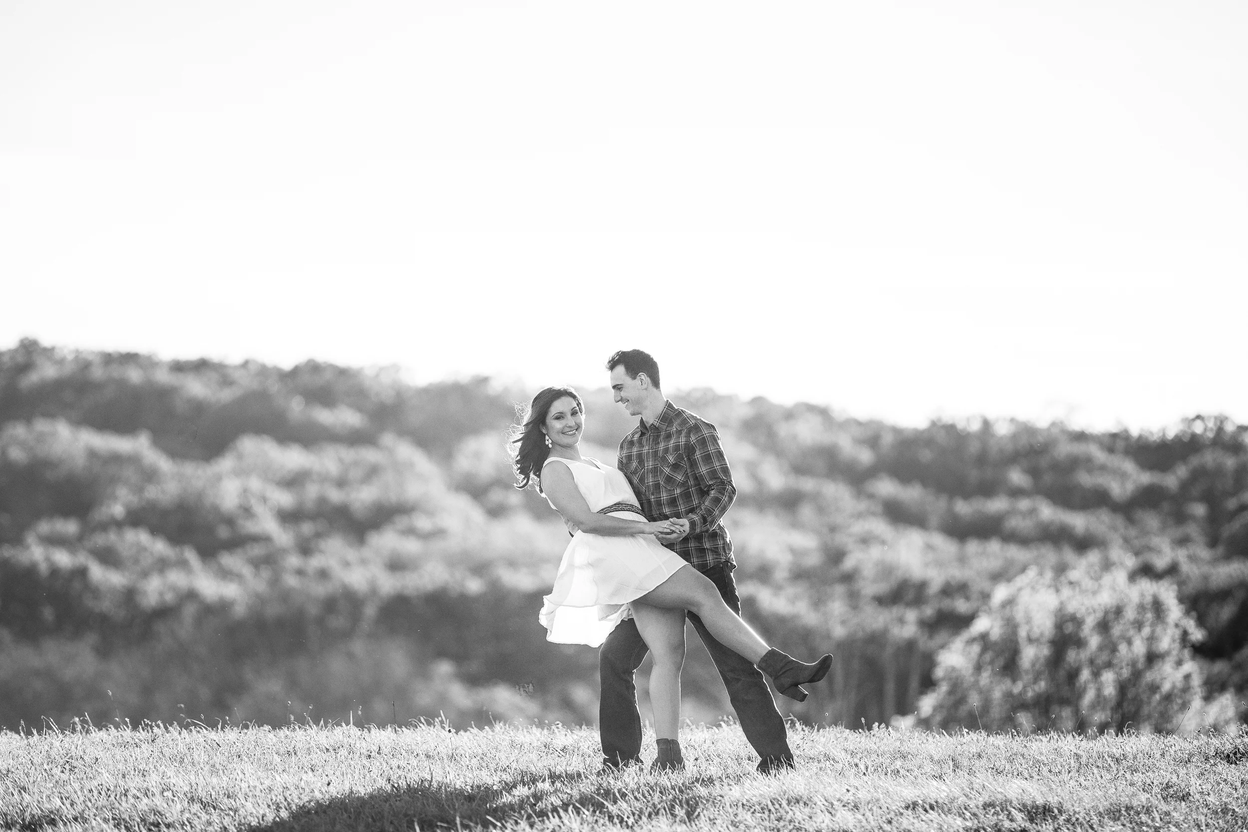 A black and white photo of a couple dancing outdoors on a grassy field with a scenic backdrop of trees and hills.