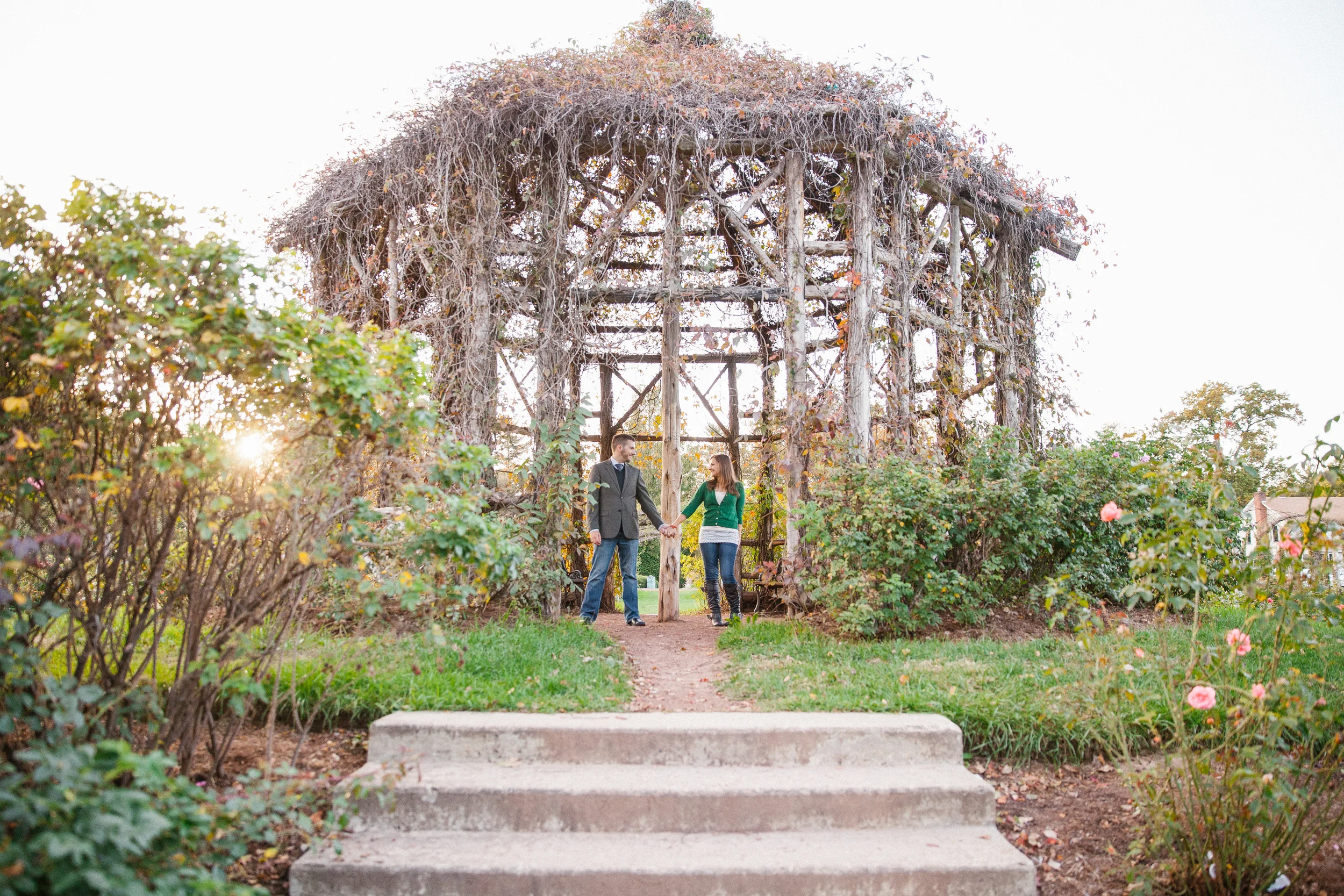 A man and woman holding hands under a wooden trellis in a garden during late afternoon.