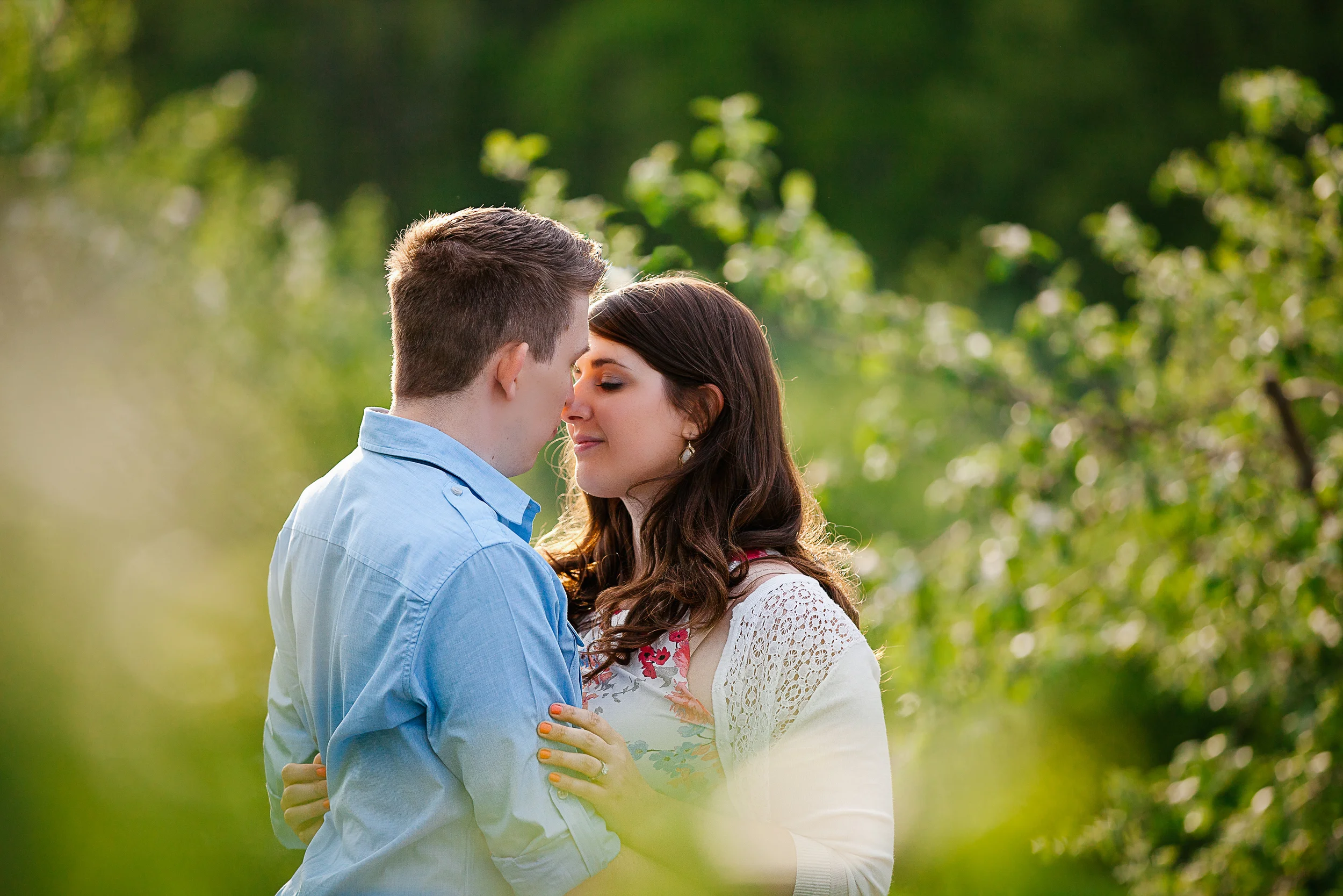 A couple standing close together outdoors with their foreheads touching, surrounded by lush green trees and soft natural lighting.