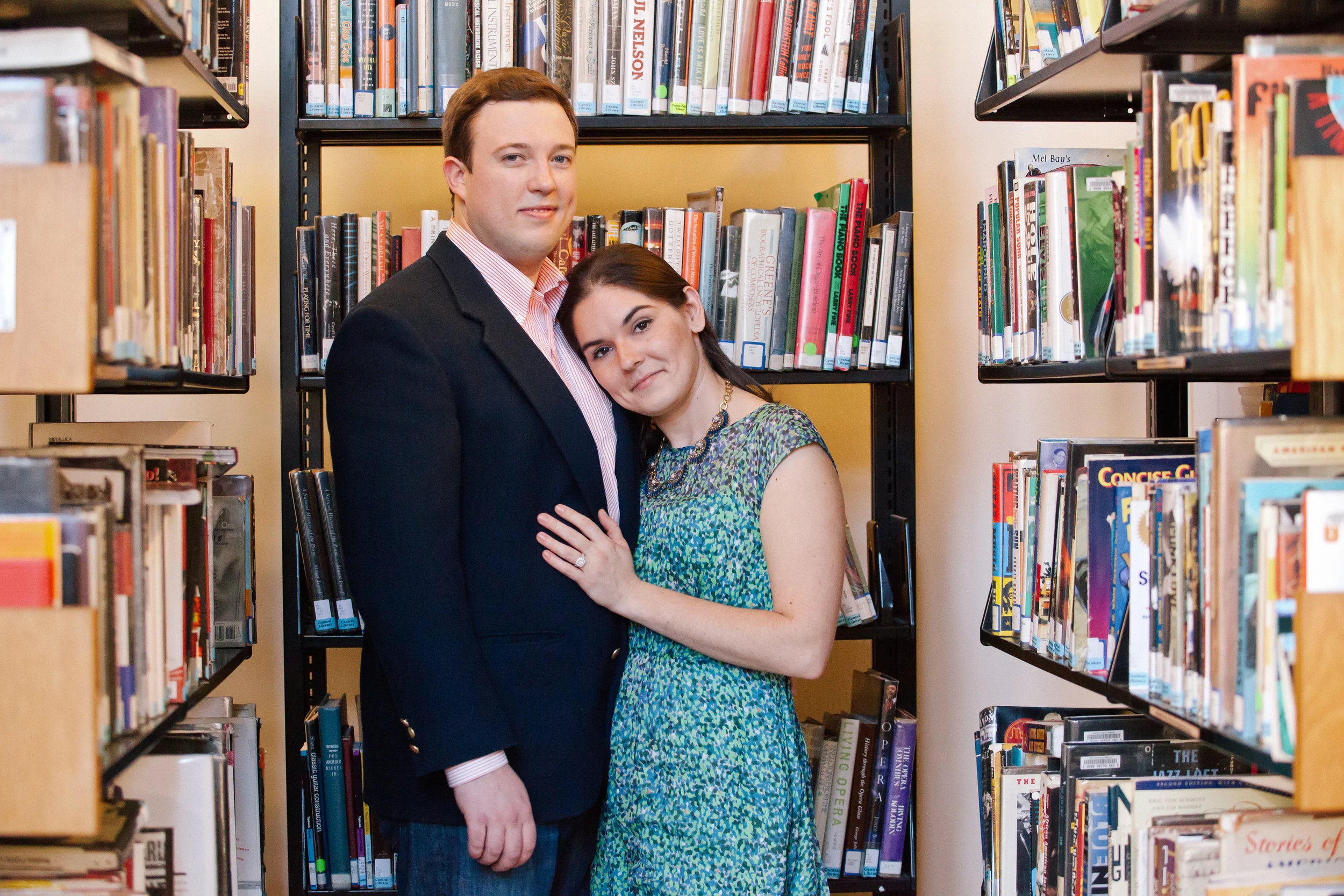 A man and woman standing closely together in a library between bookshelves, with the woman resting her head on the man's shoulder.