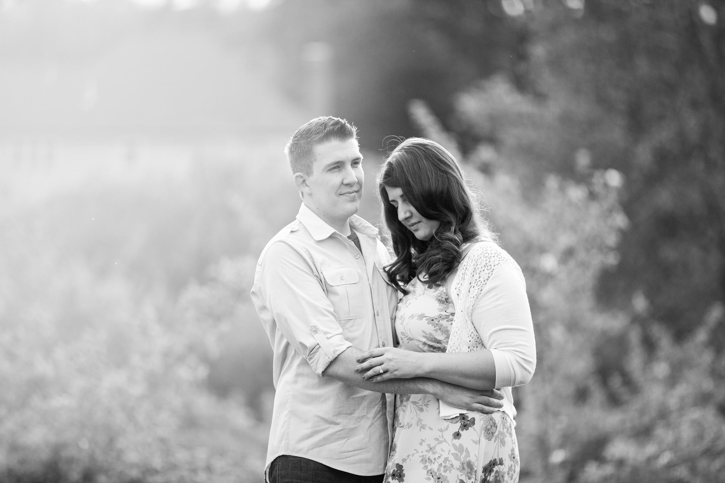 A black and white photo of a couple standing outdoors, with the man gently holding the woman's hands and the woman resting her head on his shoulder, surrounded by blurred trees and natural scenery.