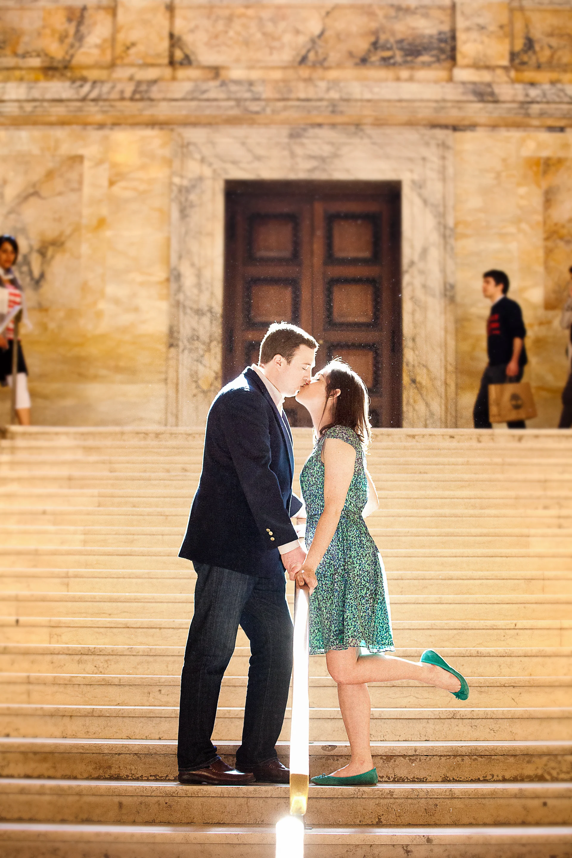 A couple stands on a staircase in front of a grand wooden door, kissing and holding hands, with two people in the background.