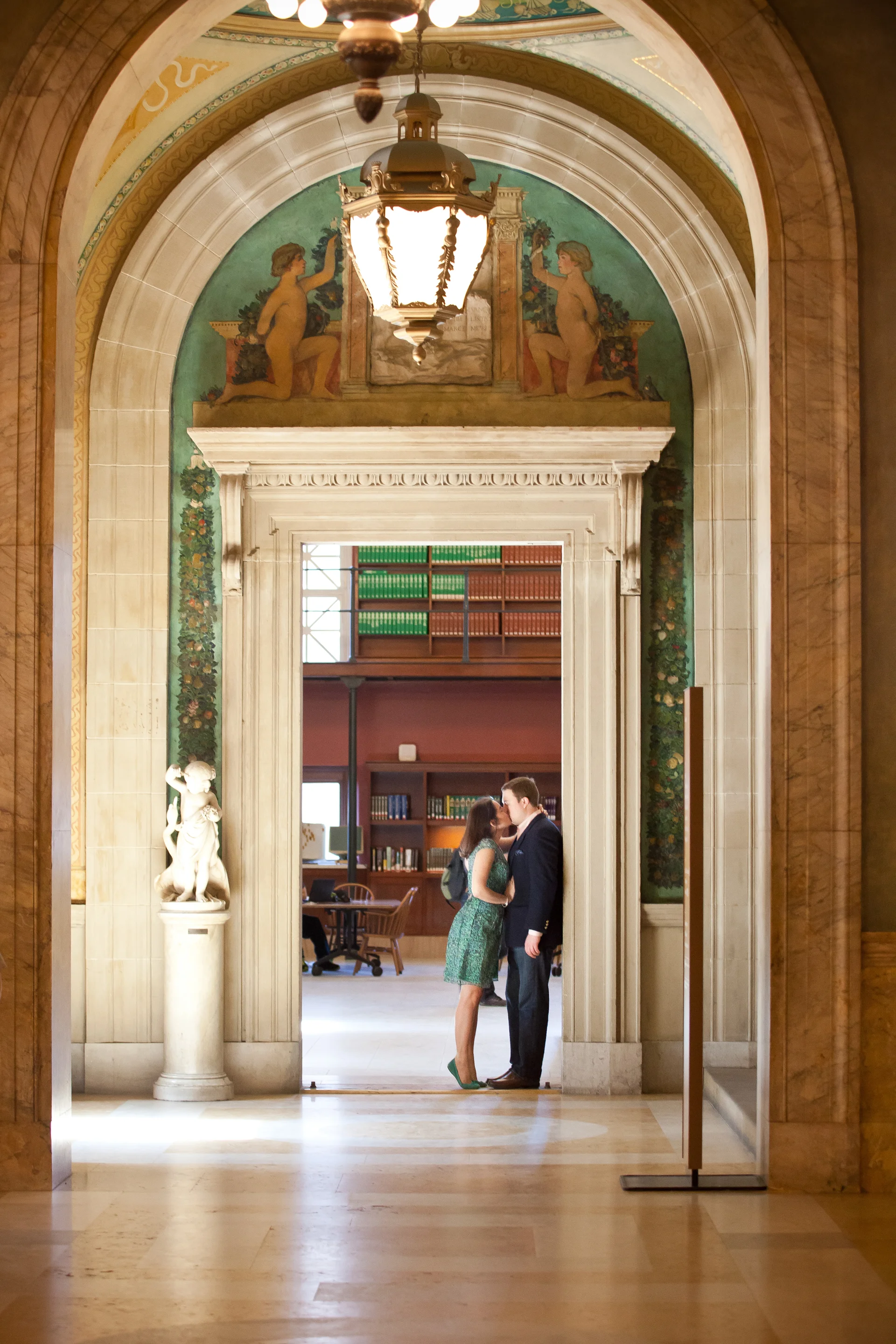 A couple sharing a kiss in a library or academic building doorway, with painted decorative art and sculptures around the entrance.