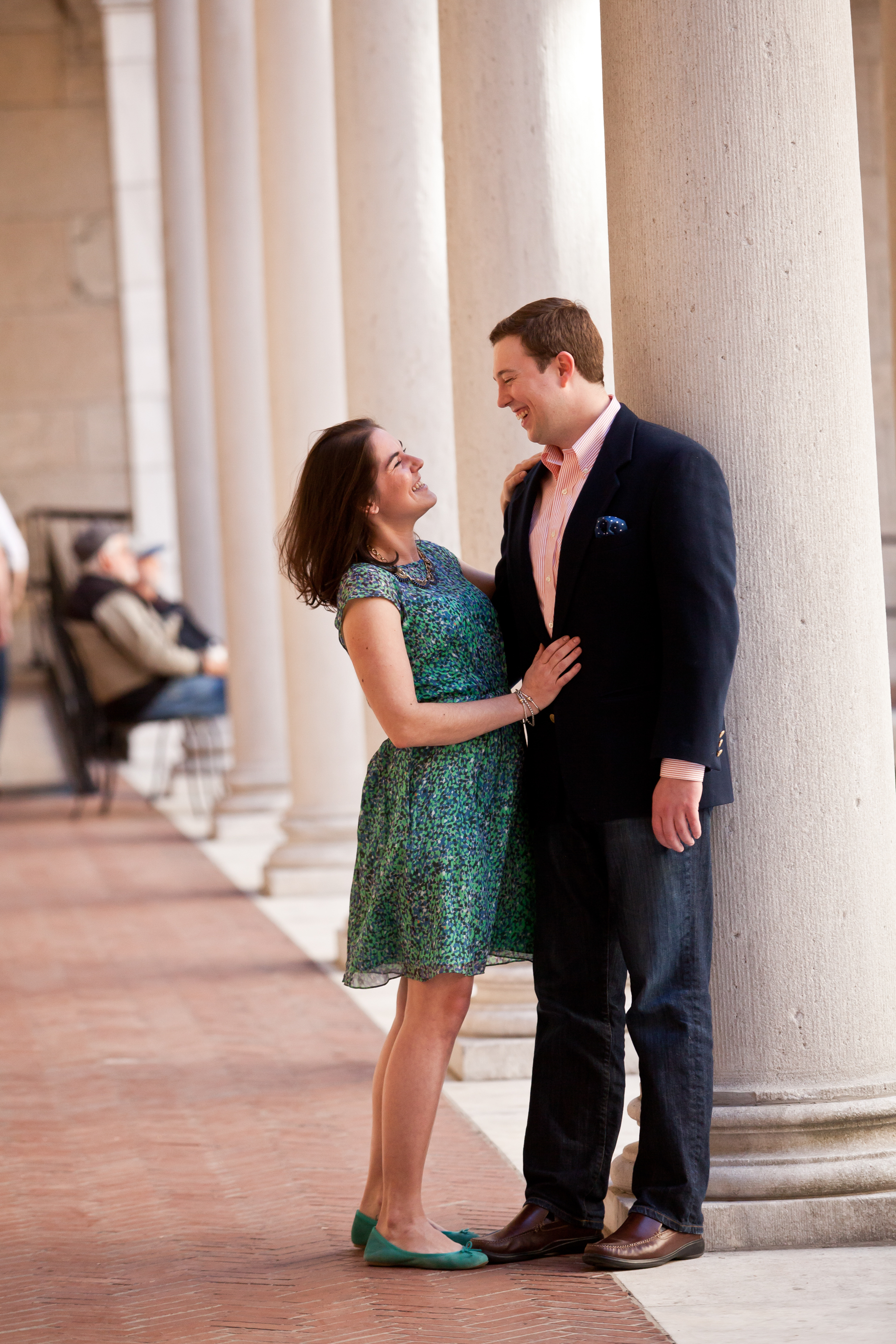 A woman and a man smiling and looking at each other, standing close against a large column in a hallway with tall columns and benches in the background.
