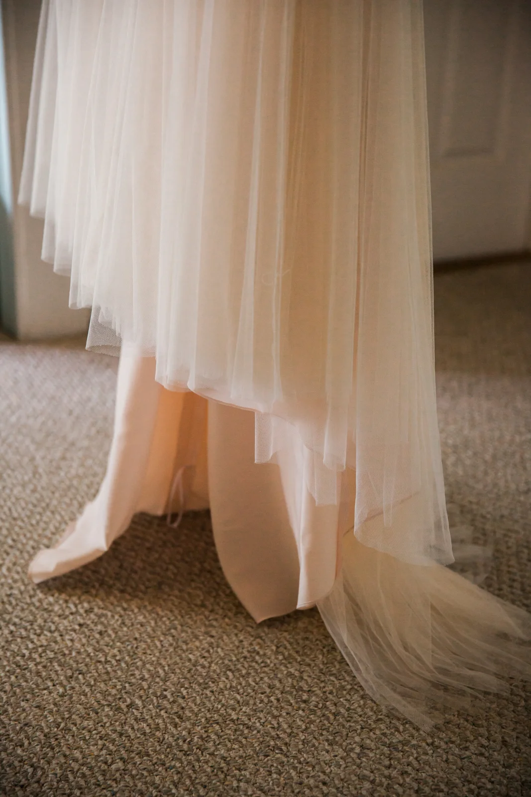Close-up of a fancy, layered, cream-colored tulle dress hanging, with the tulle fabric flowing down onto a patterned beige carpeted floor.