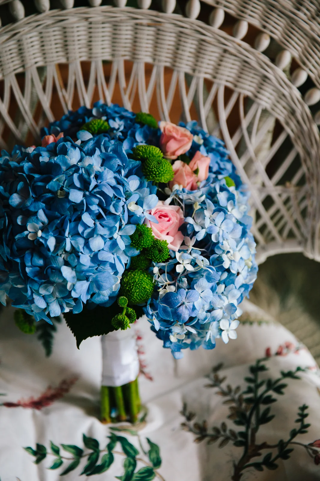 A bouquet of blue hydrangeas, pink roses, and green button poms on a chair with intricate wooden weaving, resting on a fabric with botanical embroidery.