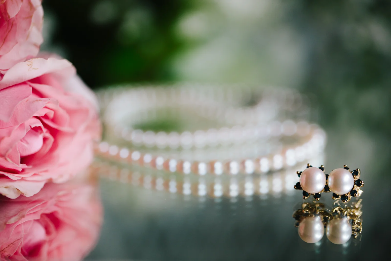 Close-up of pearl earrings with black accents, a pearl necklace in the background, and pink roses on the left side, all reflected on a shiny surface.