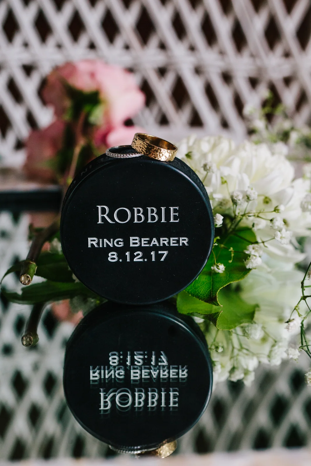 A black round case with the inscription "Robbie, Ring Bearer, 8.12.17" rests on a reflective surface, with wedding bands placed on top, surrounded by white flowers.