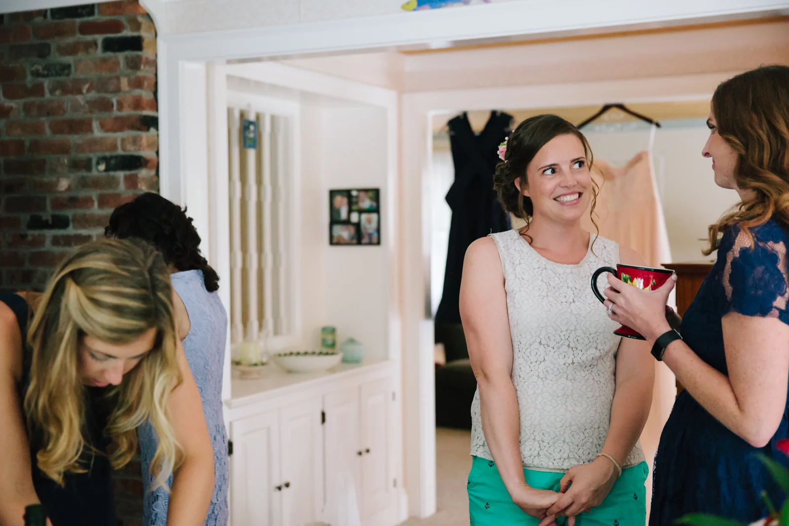 A group of women in a cozy, well-lit living room preparing for a wedding, with one woman smiling and holding a mug, and another woman showing her something inside the mug.