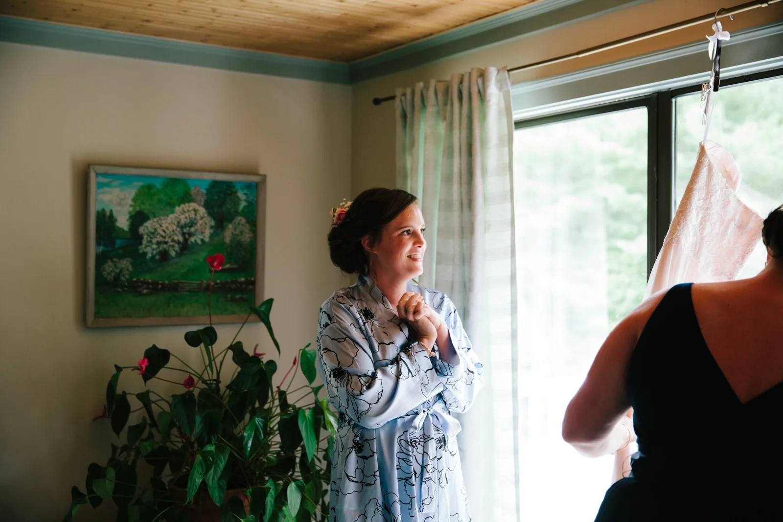 A woman in a floral robe is smiling and holding her hands together, standing near a window during daytime, with another person partially visible adjusting a pink lace garment.