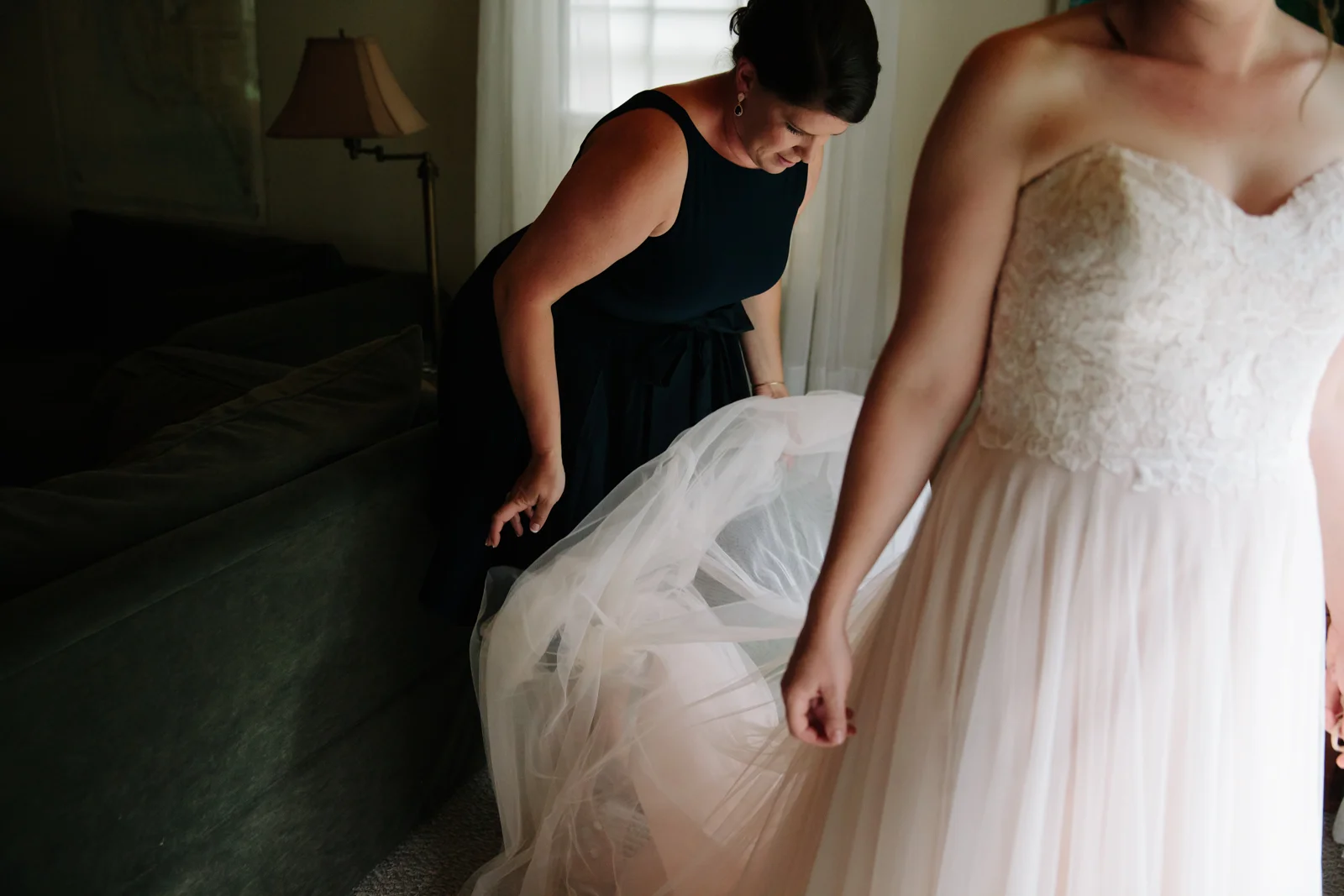 A woman in a white wedding dress standing beside another woman in a black dress who is adjusting the dress in a dimly lit room.