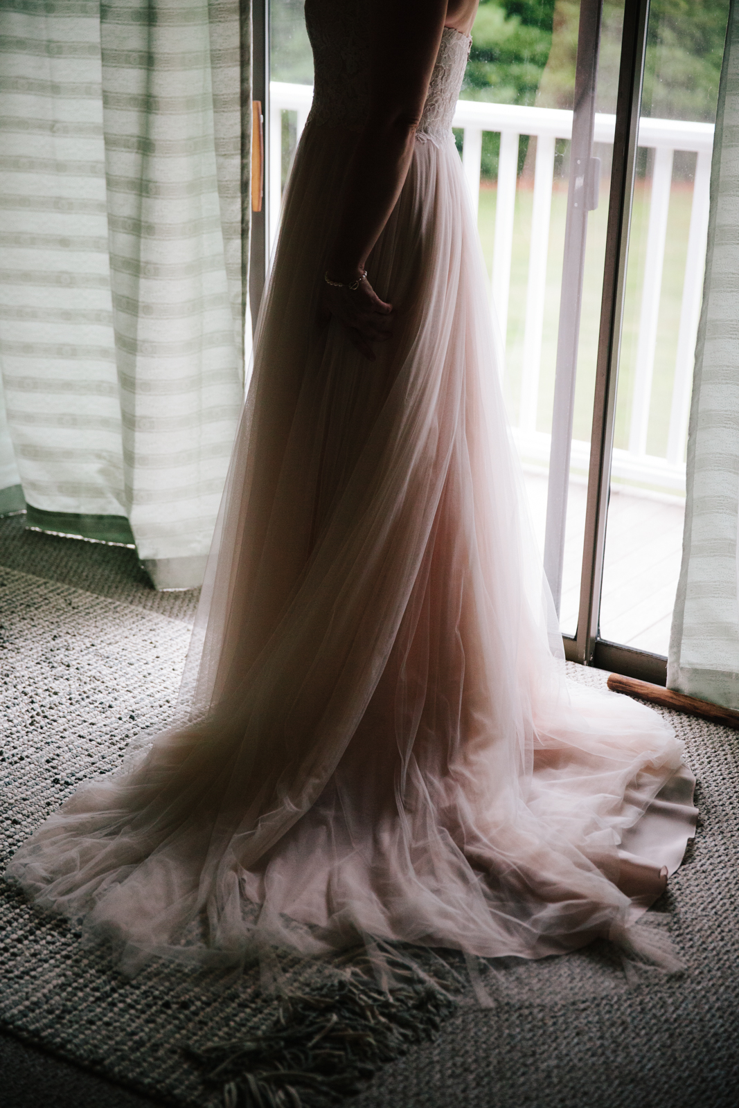 A woman in a blush pink wedding dress standing by a glass door, with her back turned, in an indoor setting.