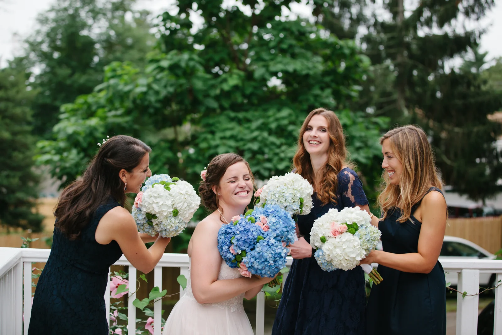 A bride and three bridesmaids celebrating outdoors, holding bouquets of white, blue, and pink flowers, with smiling faces and a wooden fence and green trees in the background.