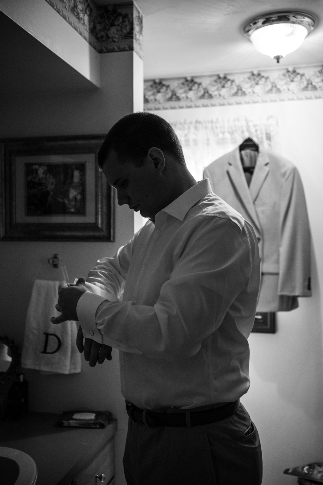 A young man in a white dress shirt is standing in a bathroom, adjusting his cufflinks, with a white jacket hanging on a hanger behind him.