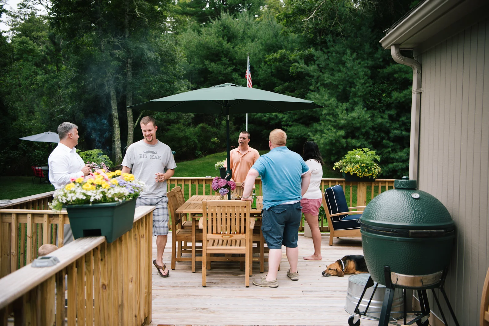 Group of five people gathered on a wooden deck outdoors, socializing near a large green barbecue grill, with flowers and patio furniture around, and trees in the background.