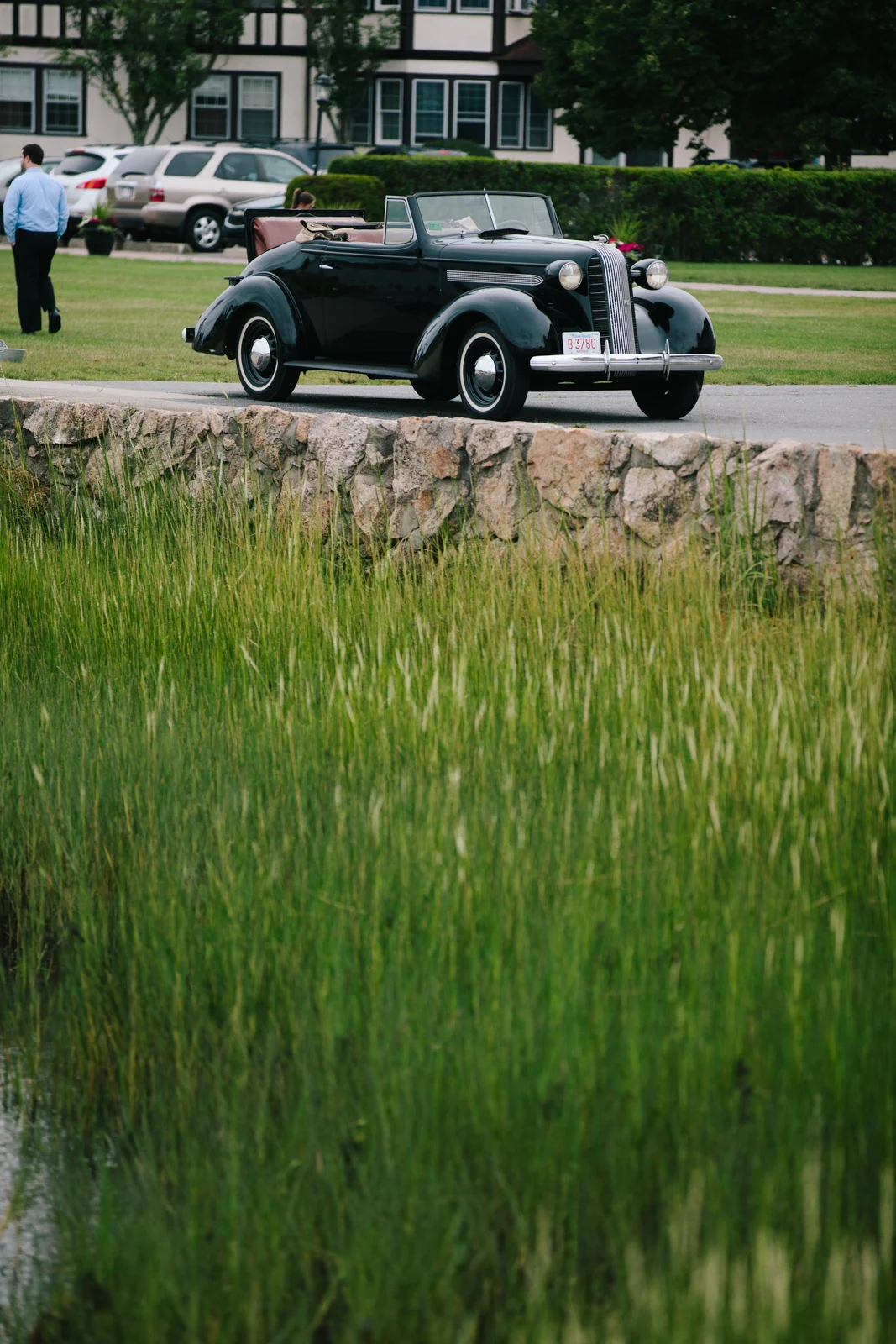A vintage black convertible car parked on a paved surface near a stone wall, with a grassy area and a building with trees in the background.