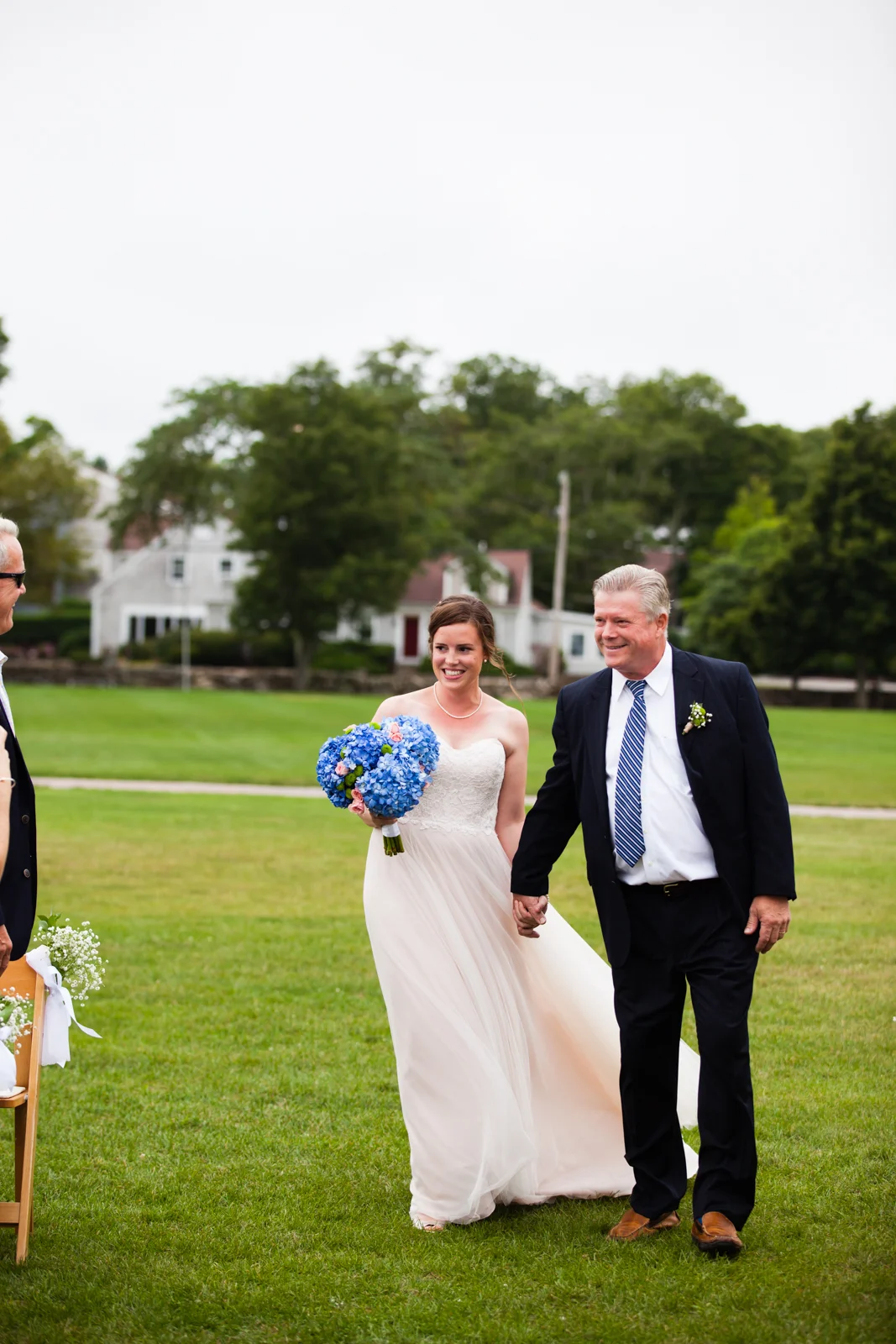 A bride holding a blue bouquet of flowers, walking outdoors with a man in a suit on a grassy field during a wedding ceremony.