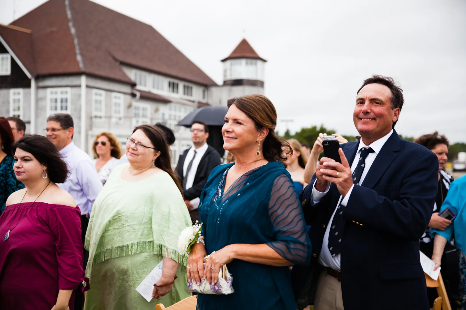 Group of people attending an outdoor formal event or wedding, with a large building that has a tower in the background.