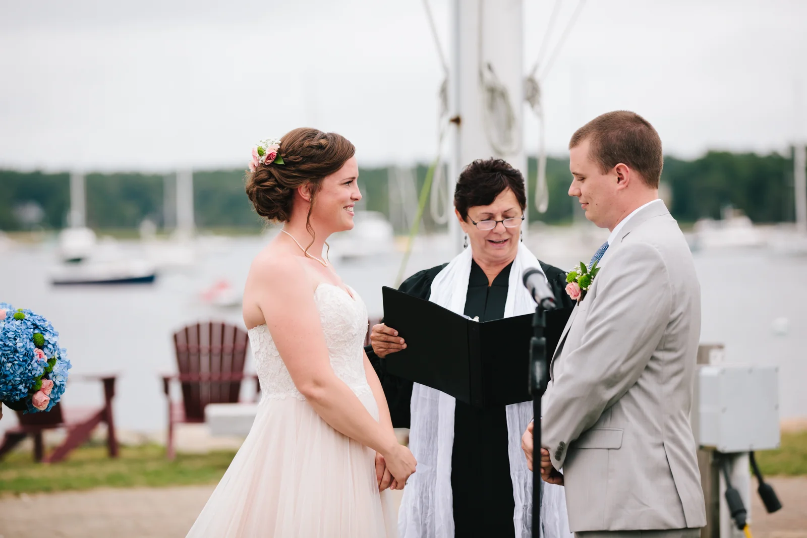 A bride and groom exchanging vows outdoors by a lake, with an officiant reading from a book during their wedding ceremony.