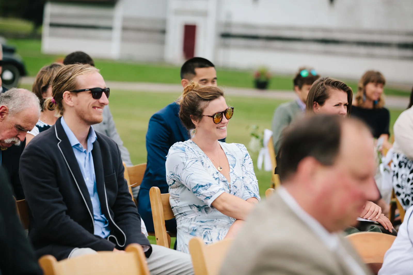 Group of people attending an outdoor event, sitting on wooden chairs on a grassy field, some smiling and wearing sunglasses.
