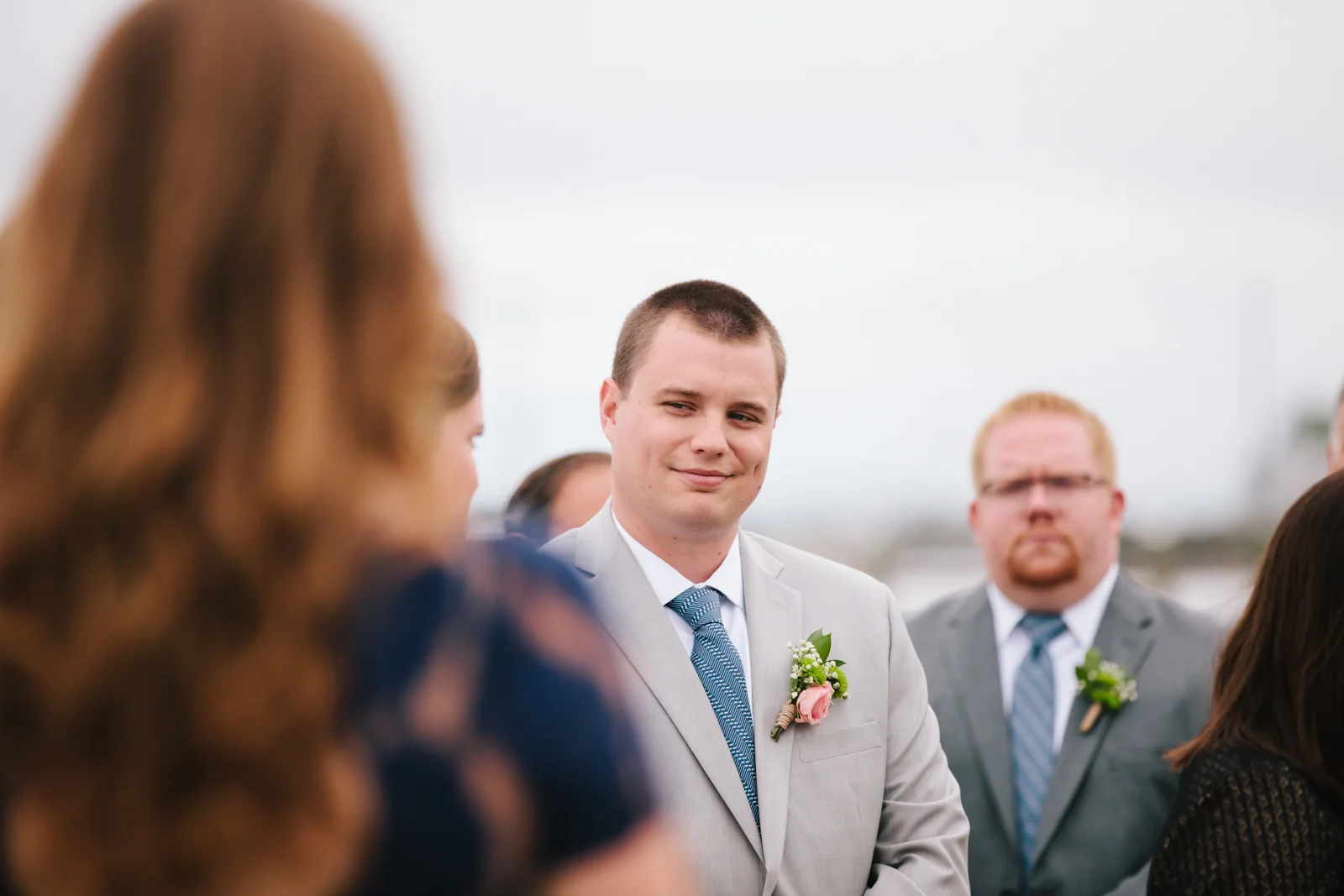 A young man in a light gray suit with a boutonniere looks at a woman with red hair during a wedding ceremony outdoors.