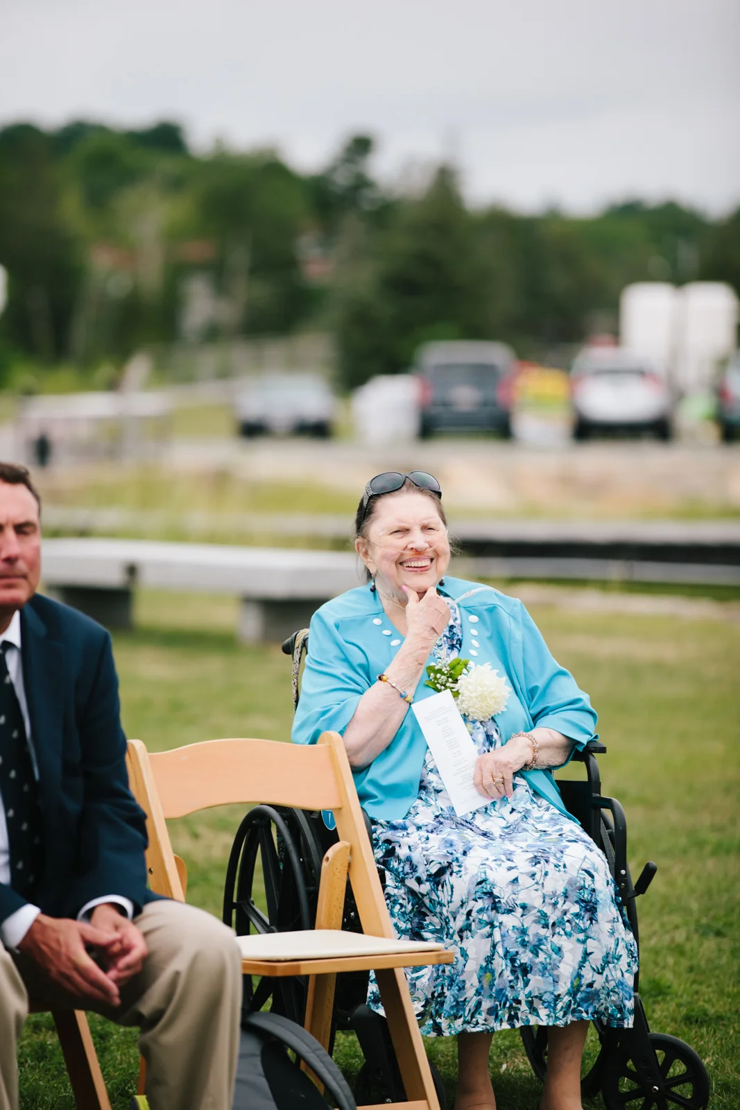An elderly woman in a wheelchair, smiling and wearing a light blue jacket with a floral dress, sitting outdoors at a gathering